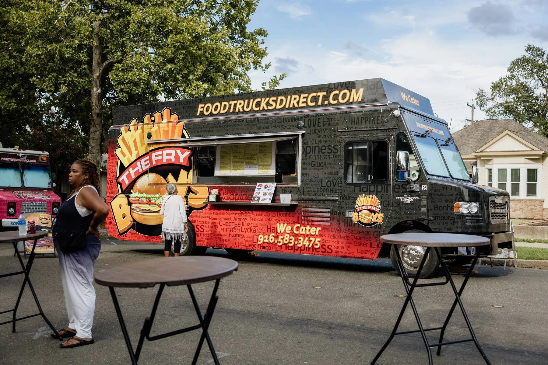 Black food truck with colorful graphics advertising burgers, with a large logo of a burger with fries and a banner for foodtrucksdirect.com, parked on a residential street with a woman and an older woman standing near it. Two black tables with chairs