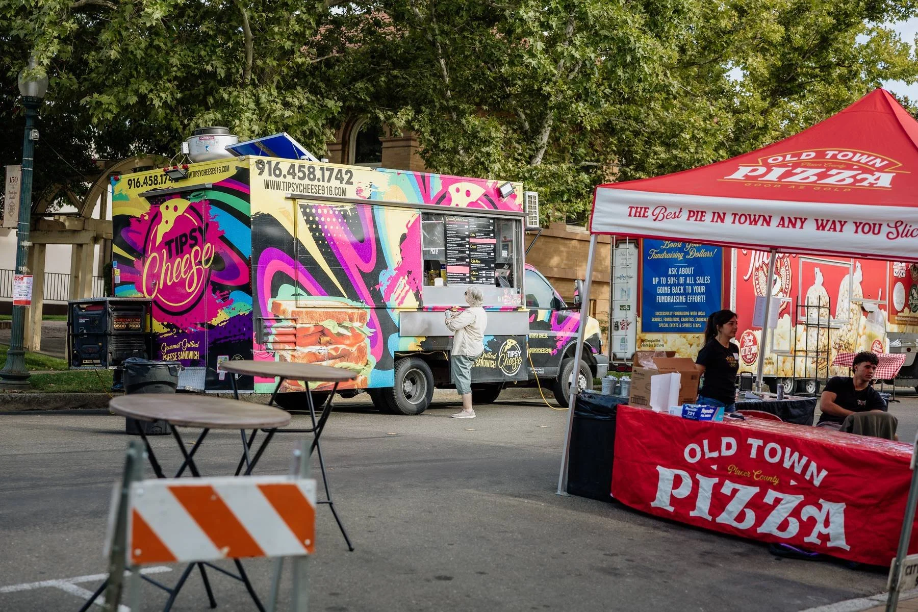 Brightly colored food truck with 'Tips Cheese' branding serving grilled cheese sandwiches, parked near a red and white 'Old Town Pizza' tent at an outdoor event, with a woman ordering and two people sitting nearby.