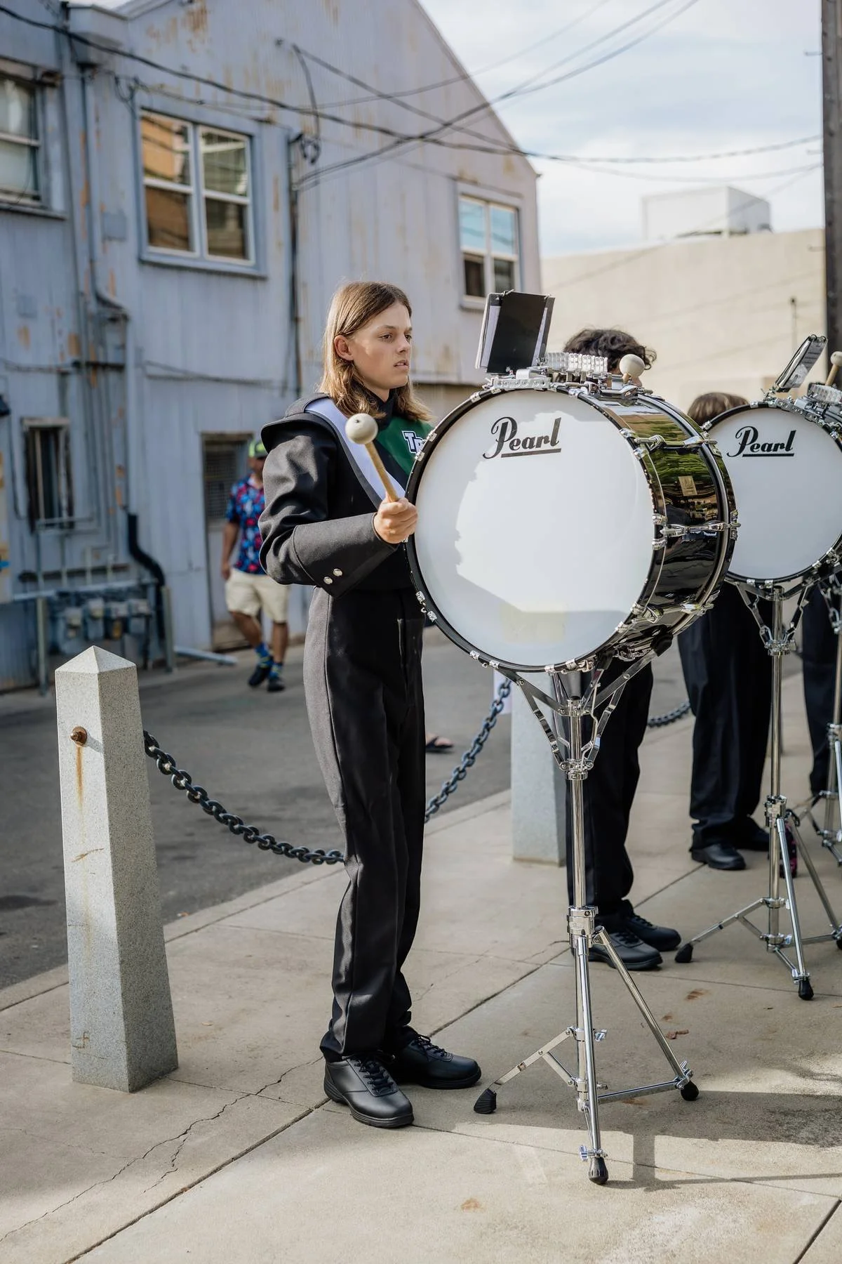 A young woman in black pants and a jacket playing a large Pearl bass drum with drumsticks outdoors, with a blue industrial building in the background.