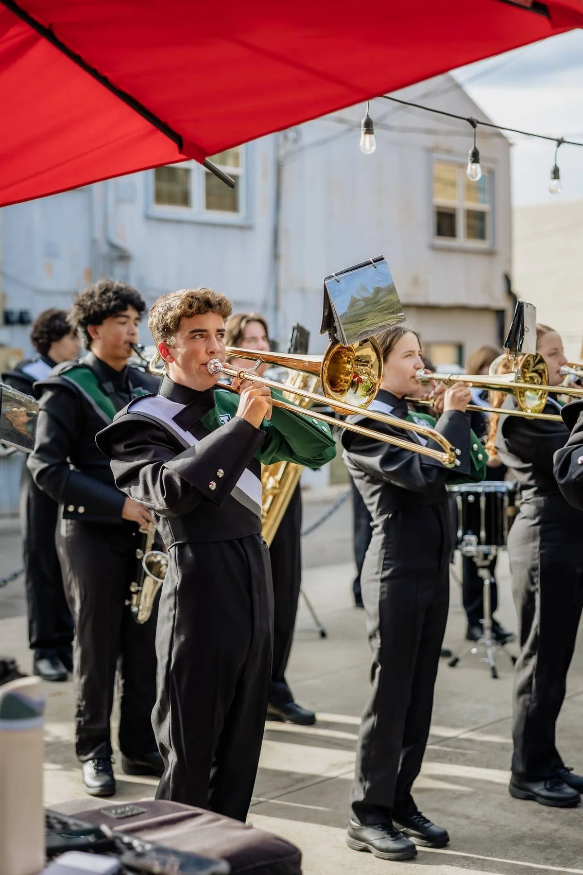 A group of young musicians playing trombones during an outdoor performance, with a red umbrella partially visible above them and a light-colored building in the background.