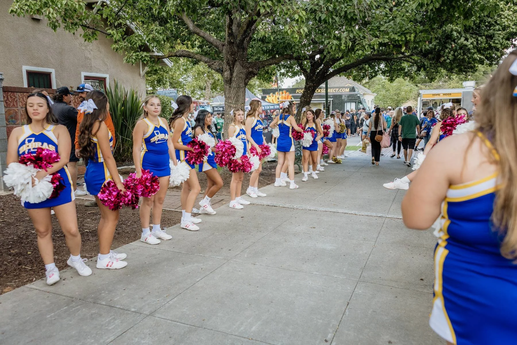 Group of cheerleaders in blue and yellow uniforms holding pink and white pom-poms standing in line outdoors near a sidewalk with trees and food trucks in the background.