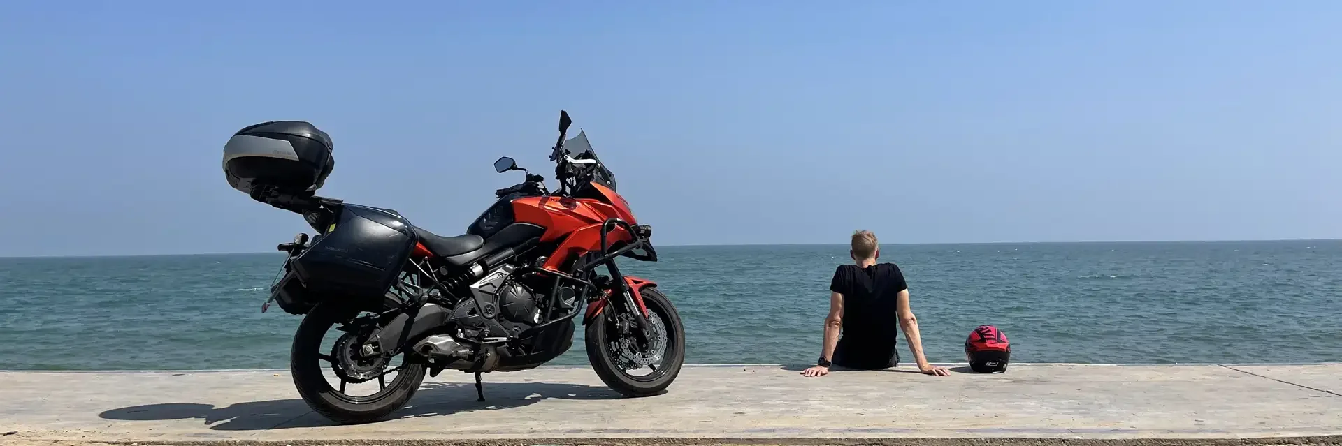 A person sitting on a concrete surface by the ocean, with a red and black motorcycle and a pink helmet nearby.