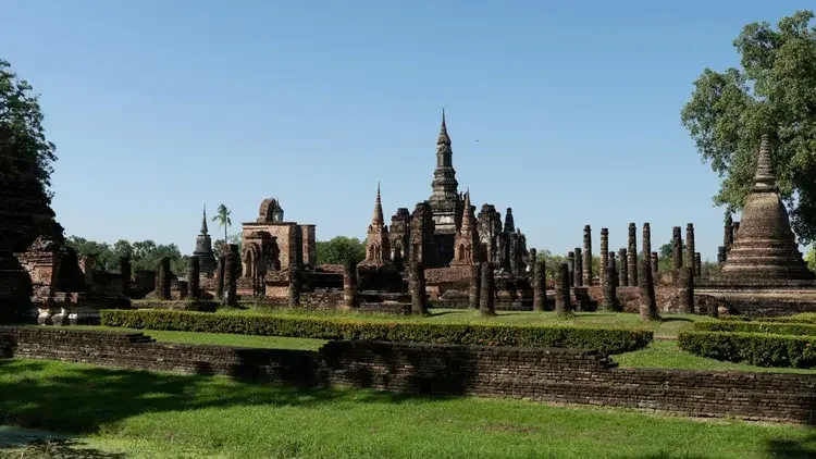Ancient Thai temple ruins with multiple spires and brick structures, surrounded by green grass and trees under a clear blue sky in Sukhothai, Thailand