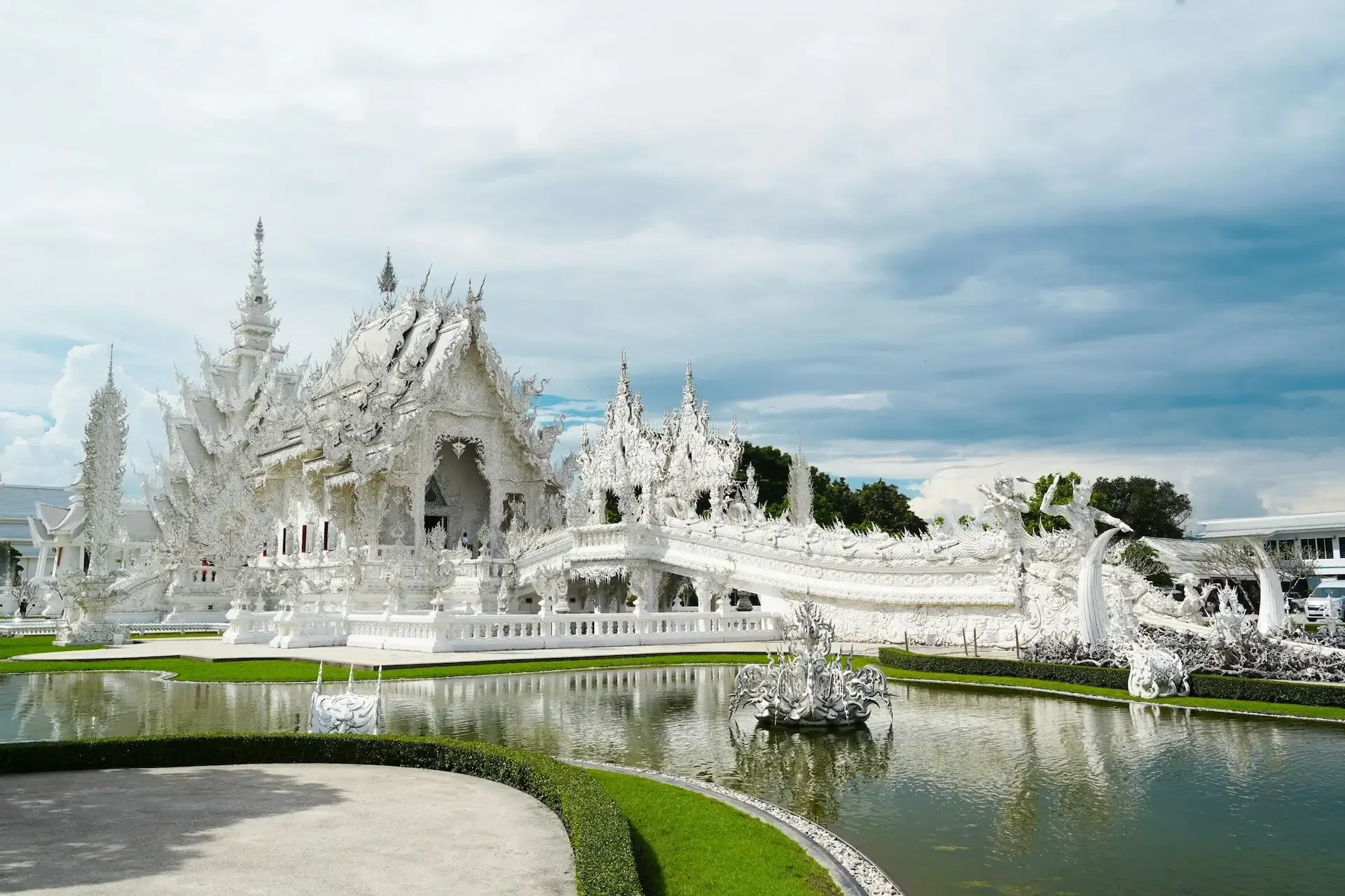 Chiang Rai white temple with intricate details, surrounded by a pond and greenery, under a cloudy sky.