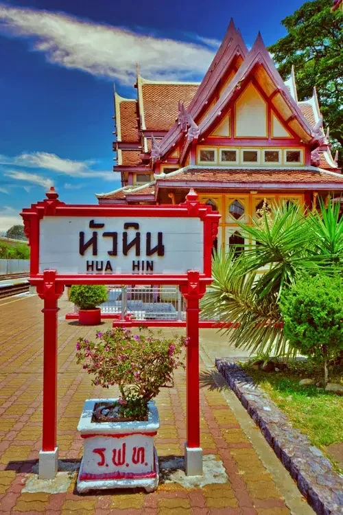 Traditional Thai-style building with ornate roof design, partly obscured by greenery. A sign in Thai and English reads 'Hua Hin' in front of the building, surrounded by flowers and plants, on a sunny day.