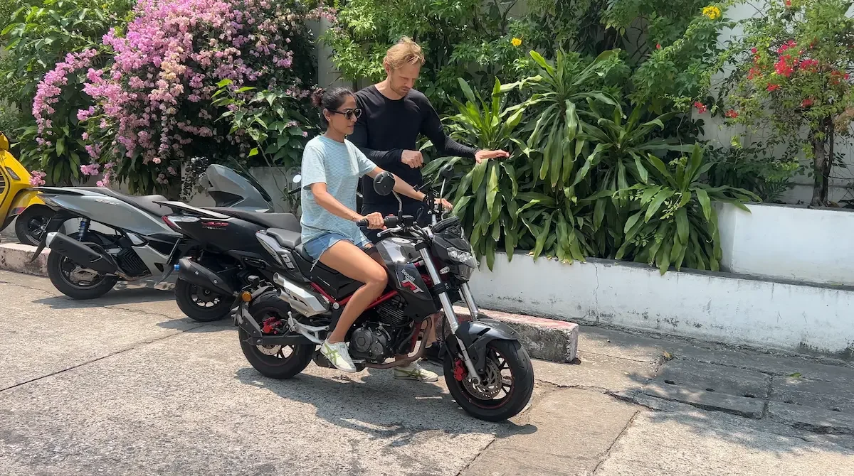 A lady riding on a small black motorbike, receiving instruction from a man standing next to her