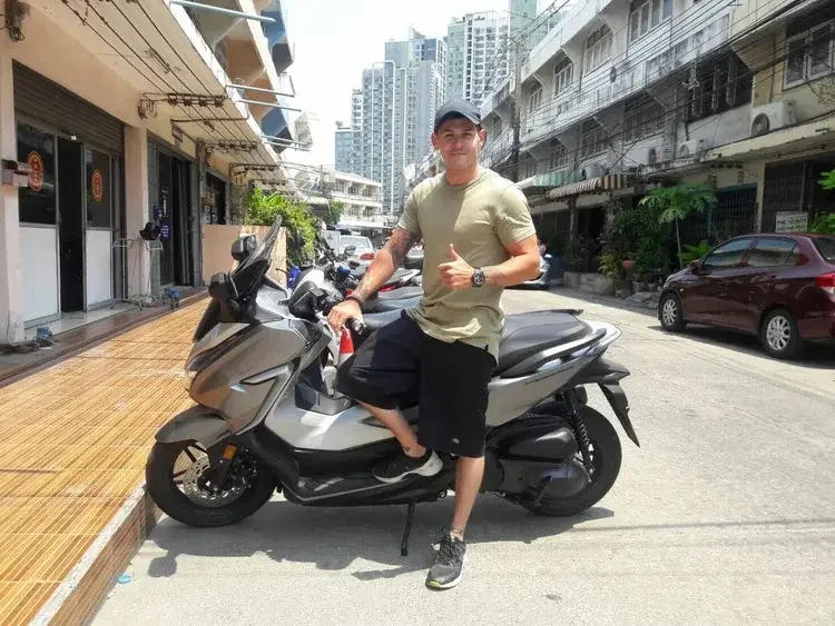 A young man in casual clothing sitting on a silver scooter in front of BSR Bike Shop in Bangkok, giving a thumbs-up.