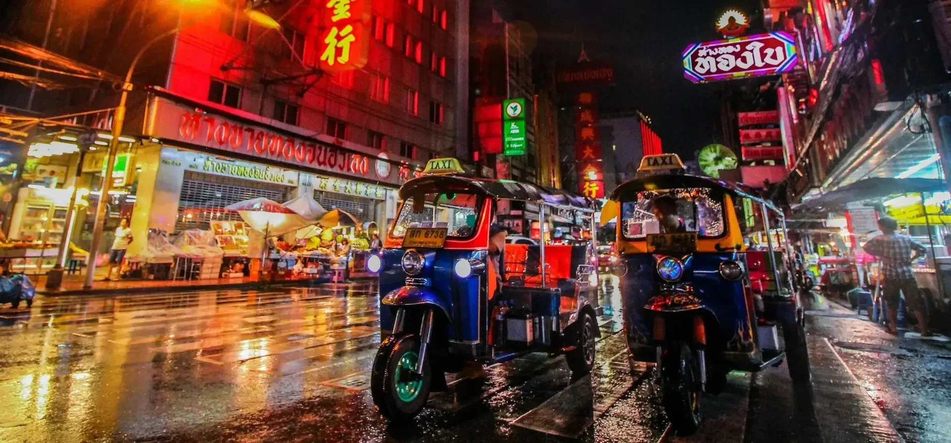 Street scene at night with colorful neon signs, two tuk-tuks on a wet road, and street vendors.