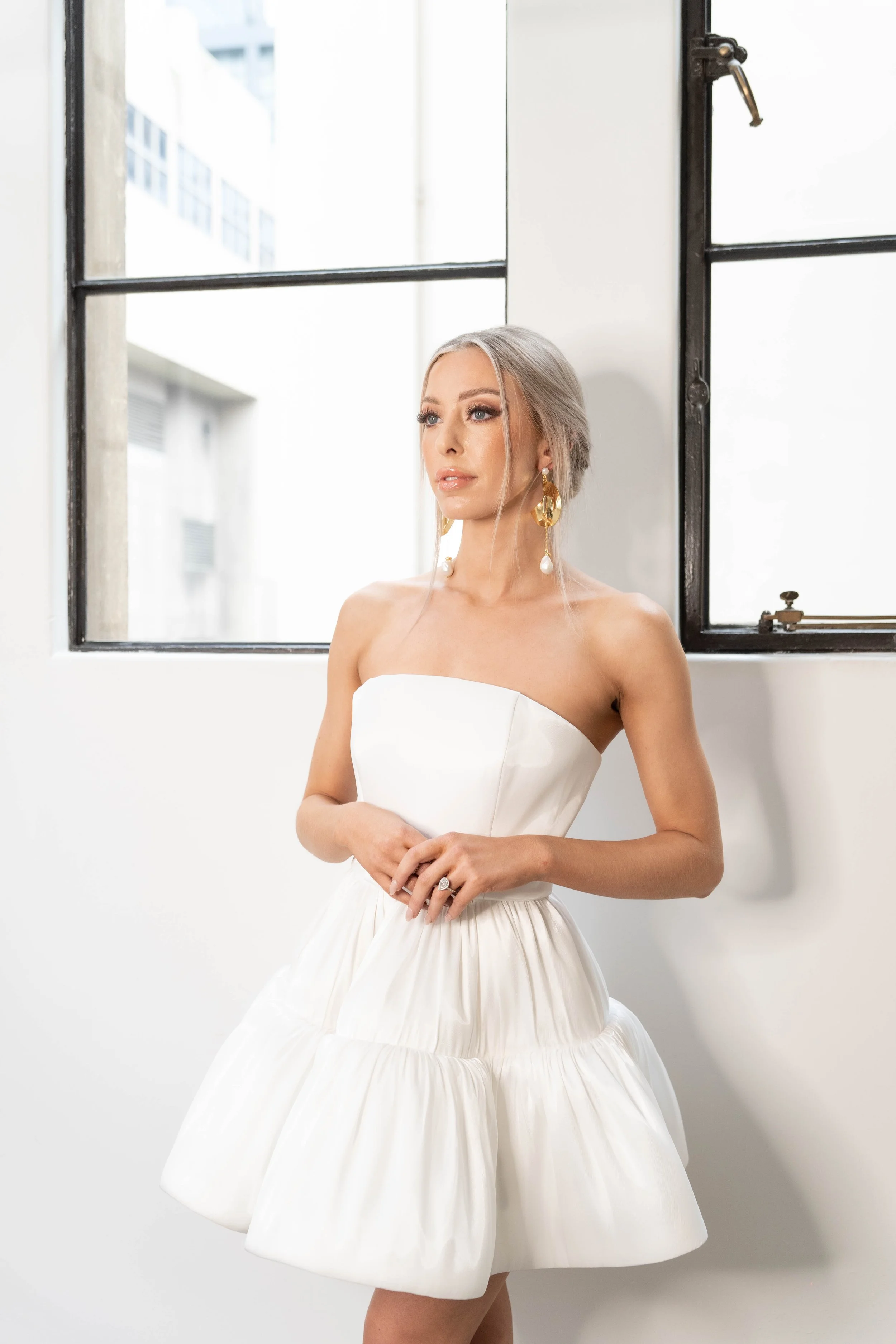 A woman in a strapless white dress standing in front of a window with metal framing, looking to the side.