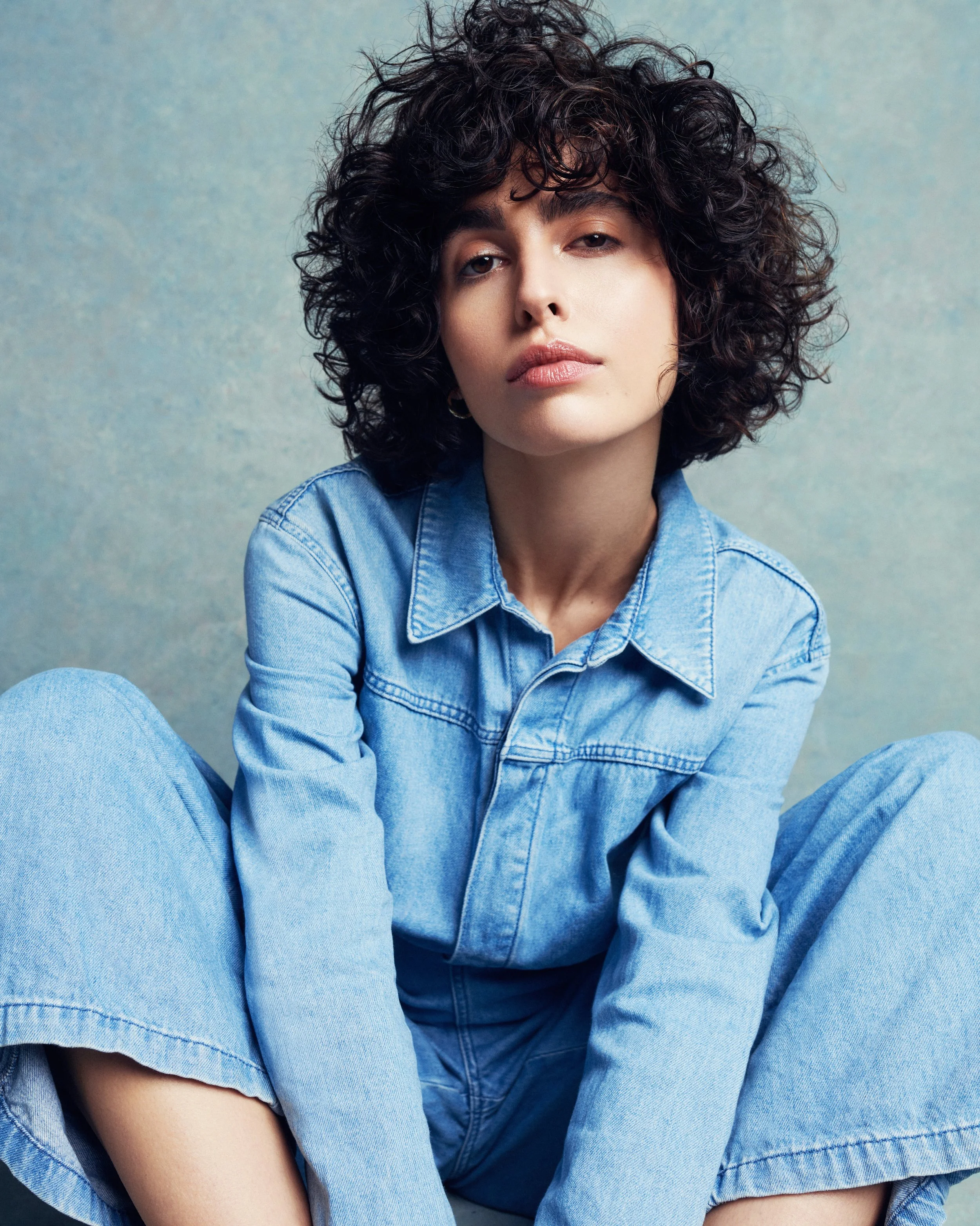 Close-up portrait of a woman with curly dark hair, wearing a blue denim shirt, against a muted blue background.