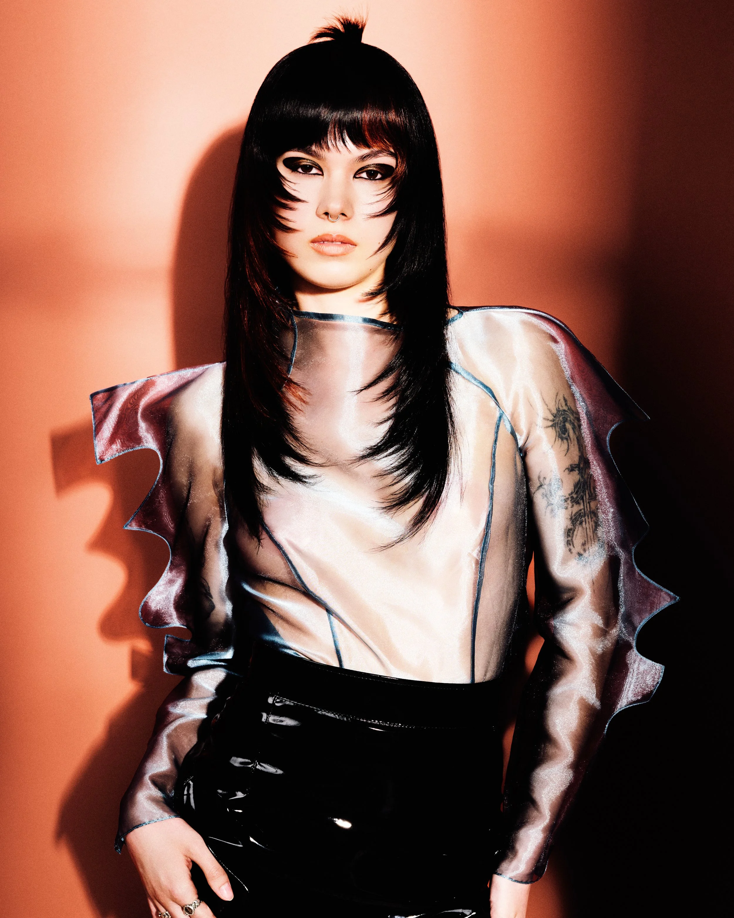 A woman with black hair, makeup, and tattoos wearing a shiny, sheer top, posing against a pink background with shadows.