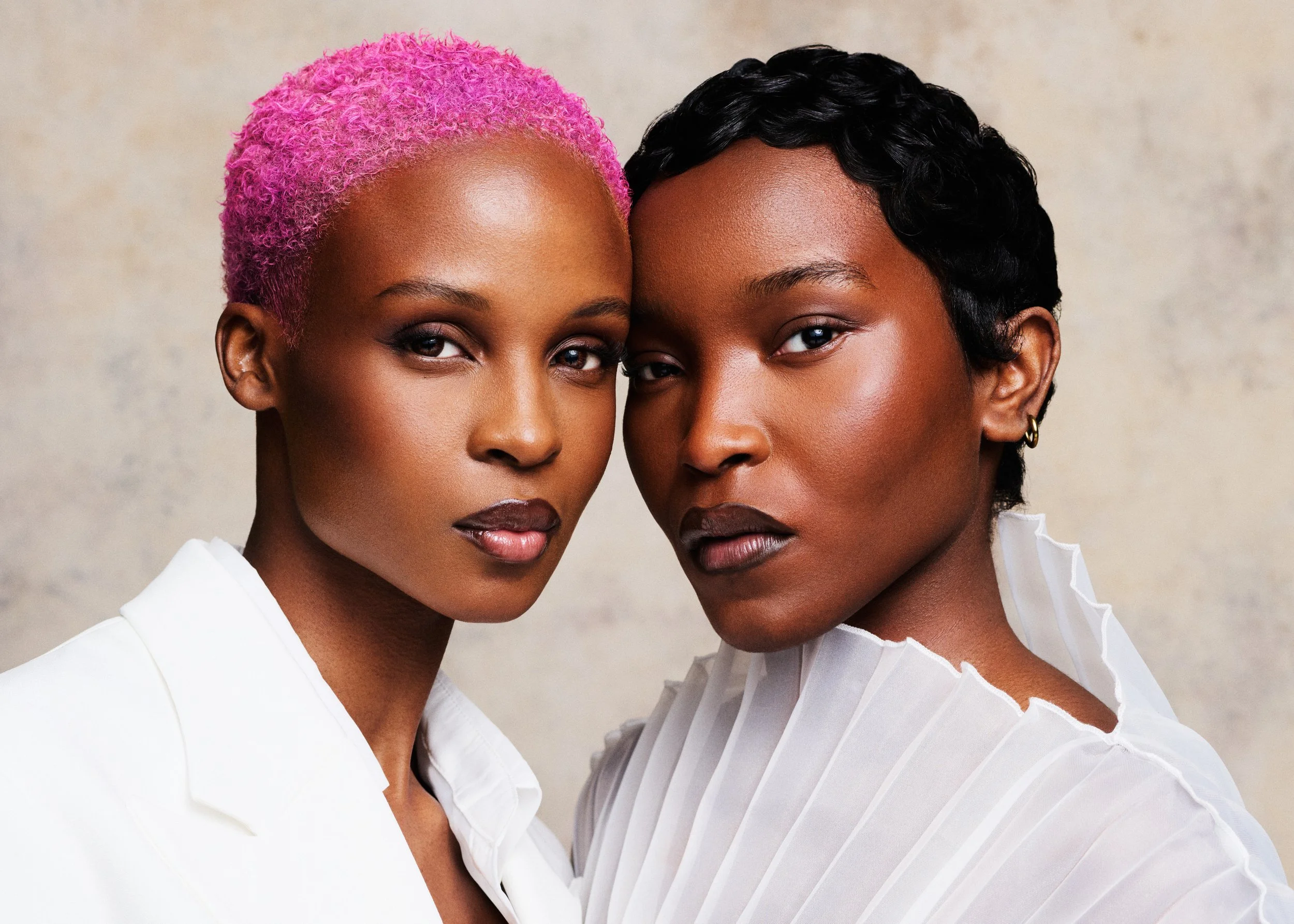 Two women with short dark hair and minimal makeup posing close together, one with bright pink curly hair, both wearing white tops with ruffled collars, against a neutral background.