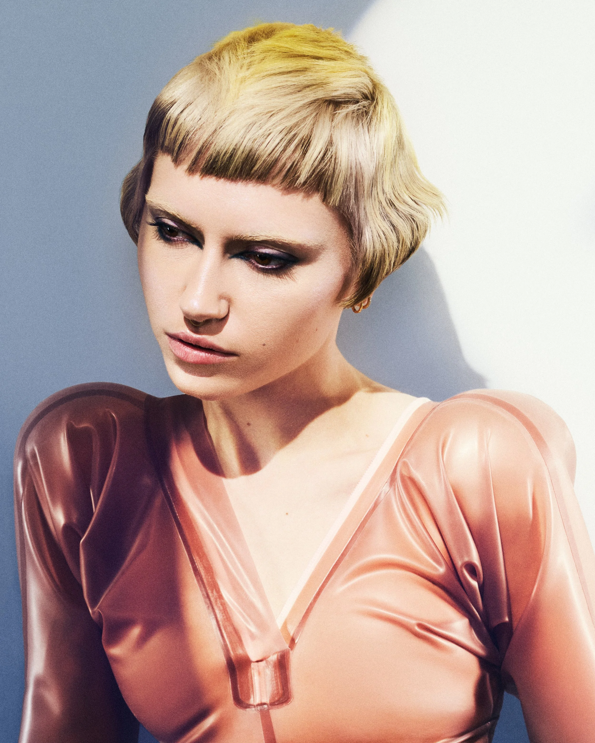 A woman with short, blond hair styled in a textured, layered cut. She is wearing makeup and a pastel-colored, shiny, translucent top.