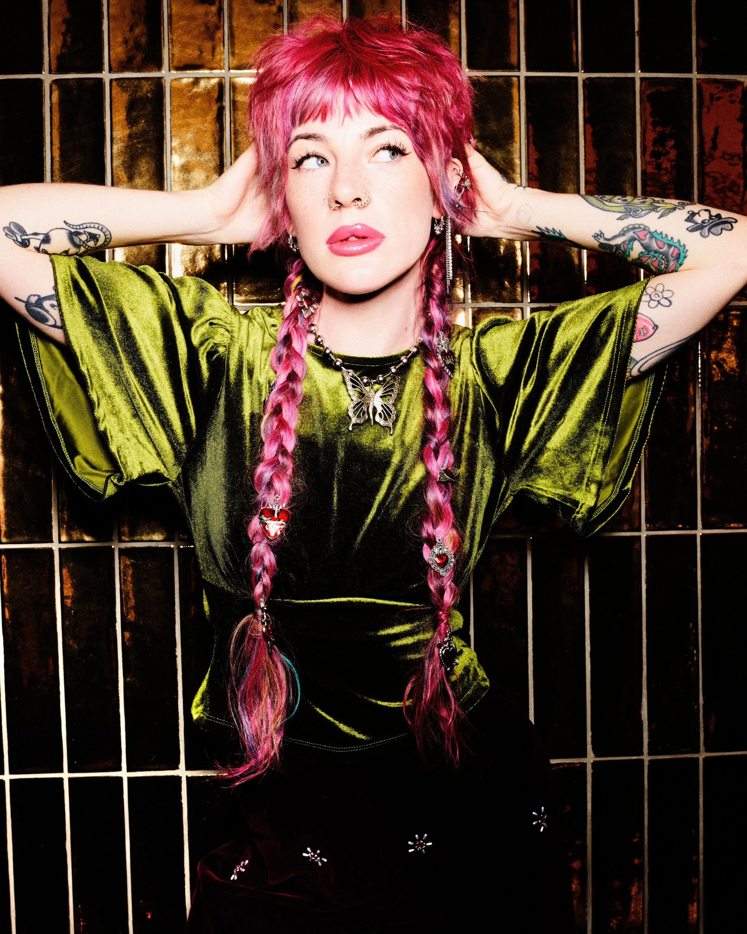 A woman with pink hair styled in braids, wearing a shiny lime green top, multiple earrings, and a butterfly necklace, stands in front of a tiled wall with metallic and dark brown tiles.