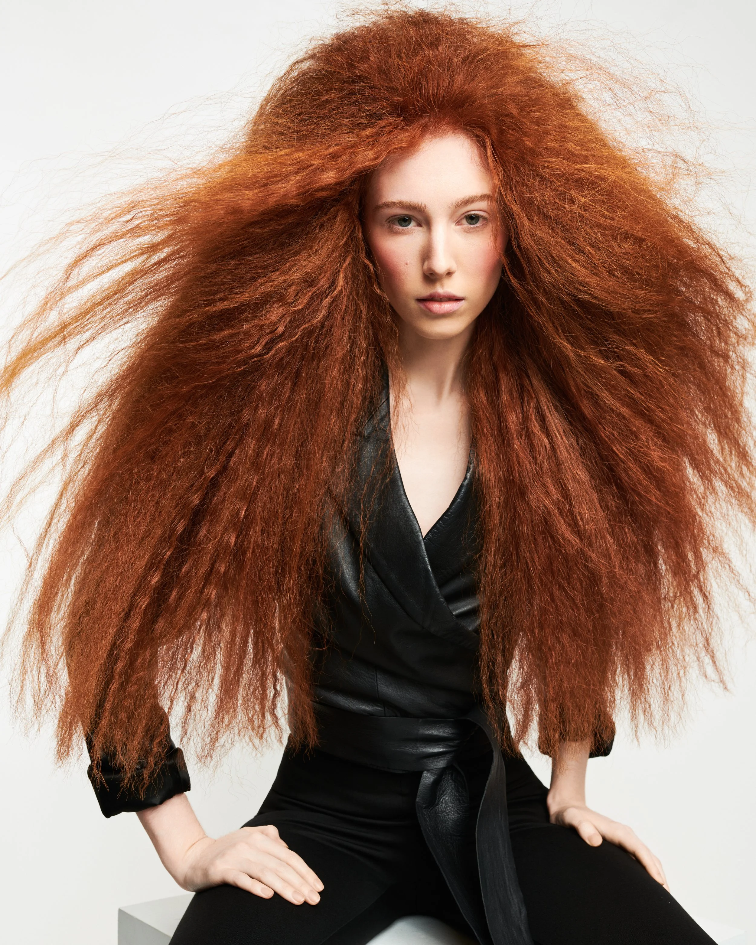 Woman with vibrant, curly red hair wearing a black outfit sitting on a white block against a plain white background.
