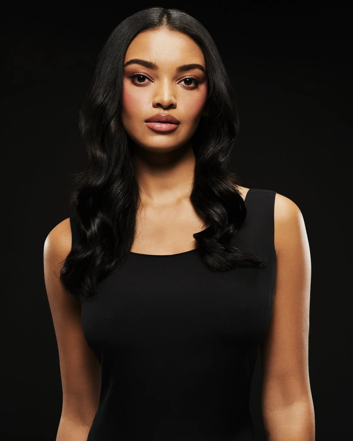 Portrait of a woman with long black hair, wearing a black sleeveless top, against a black background.