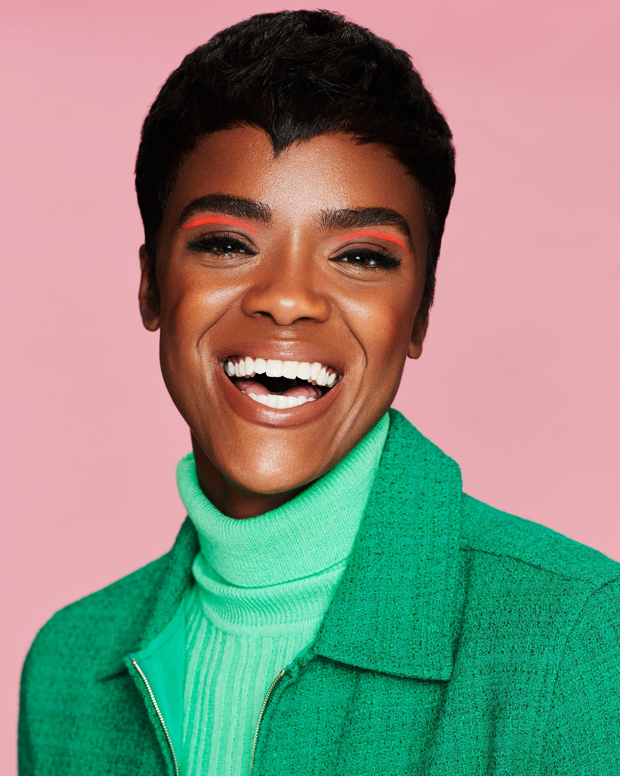 Close-up of a smiling woman with short black hair, bright makeup, wearing a green sweater against a pink background.