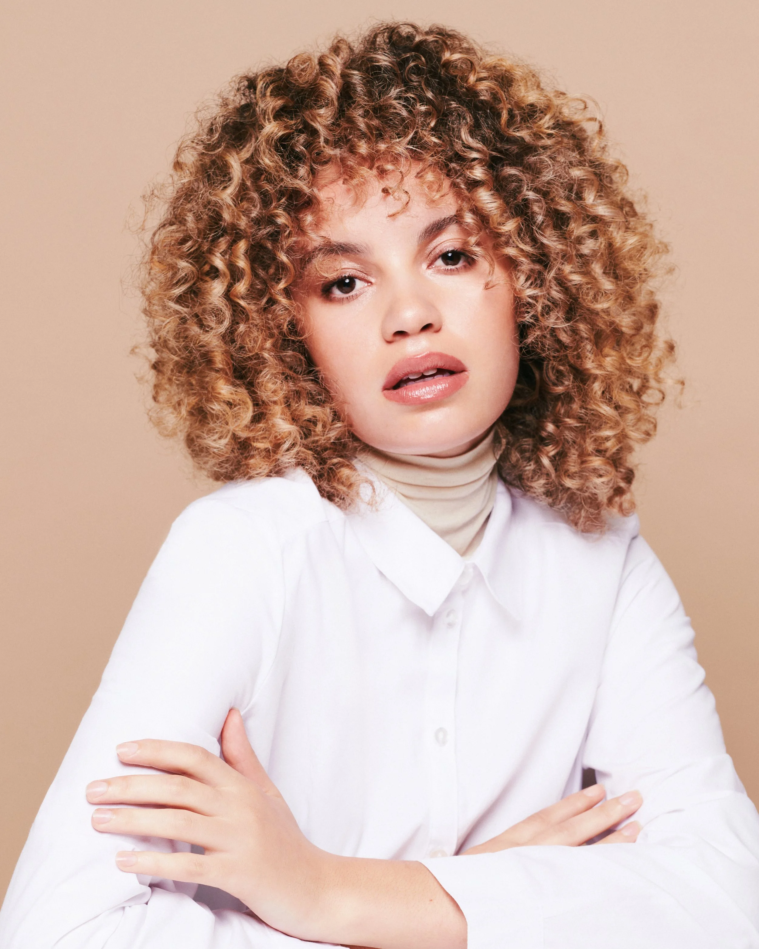 Close-up of a young woman with curly blonde hair, wearing a white shirt, with arms crossed and a neutral facial expression, standing against a beige background.