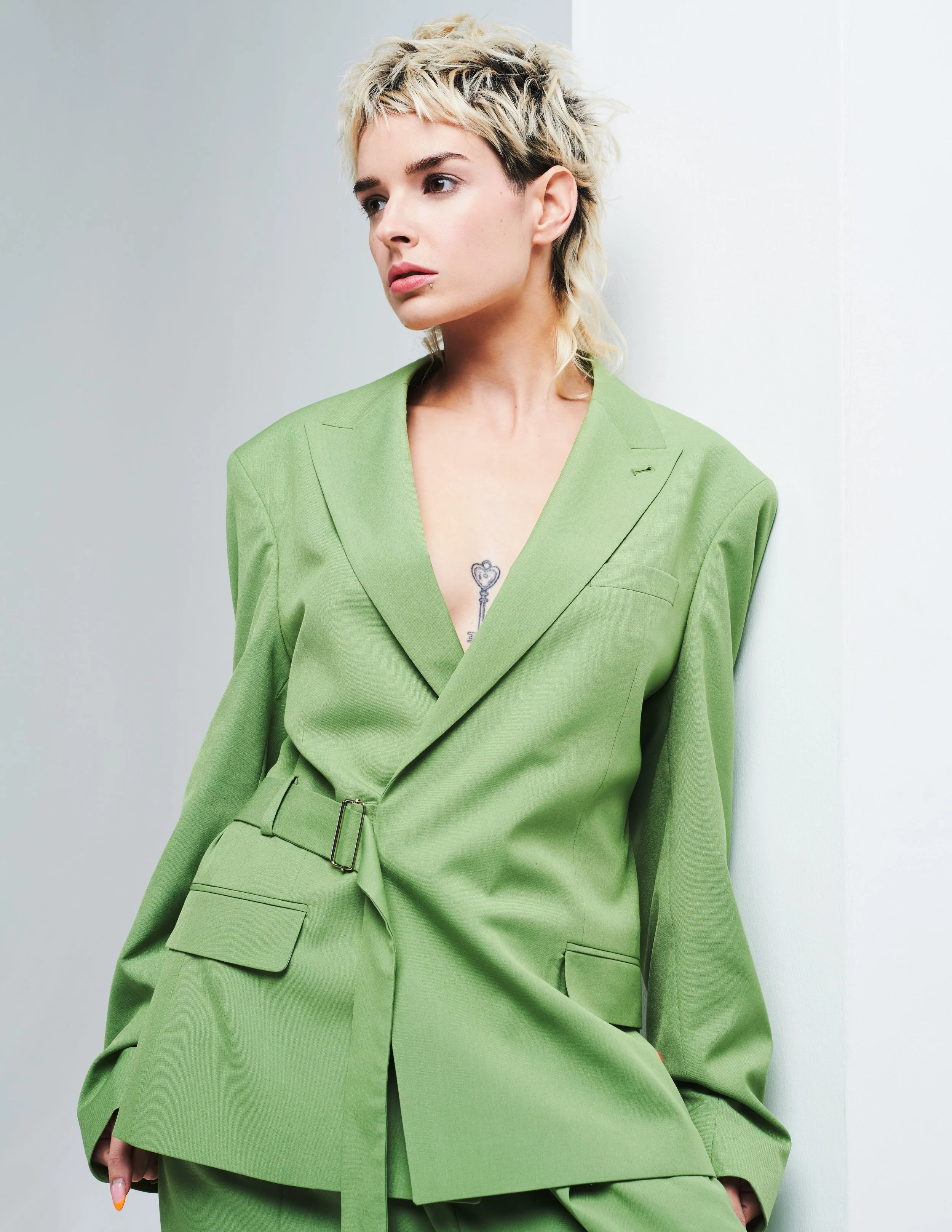 A woman with short, messy blonde hair, wearing a bright green blazer and matching pants, stands against a plain white and gray background, looking slightly to the side.