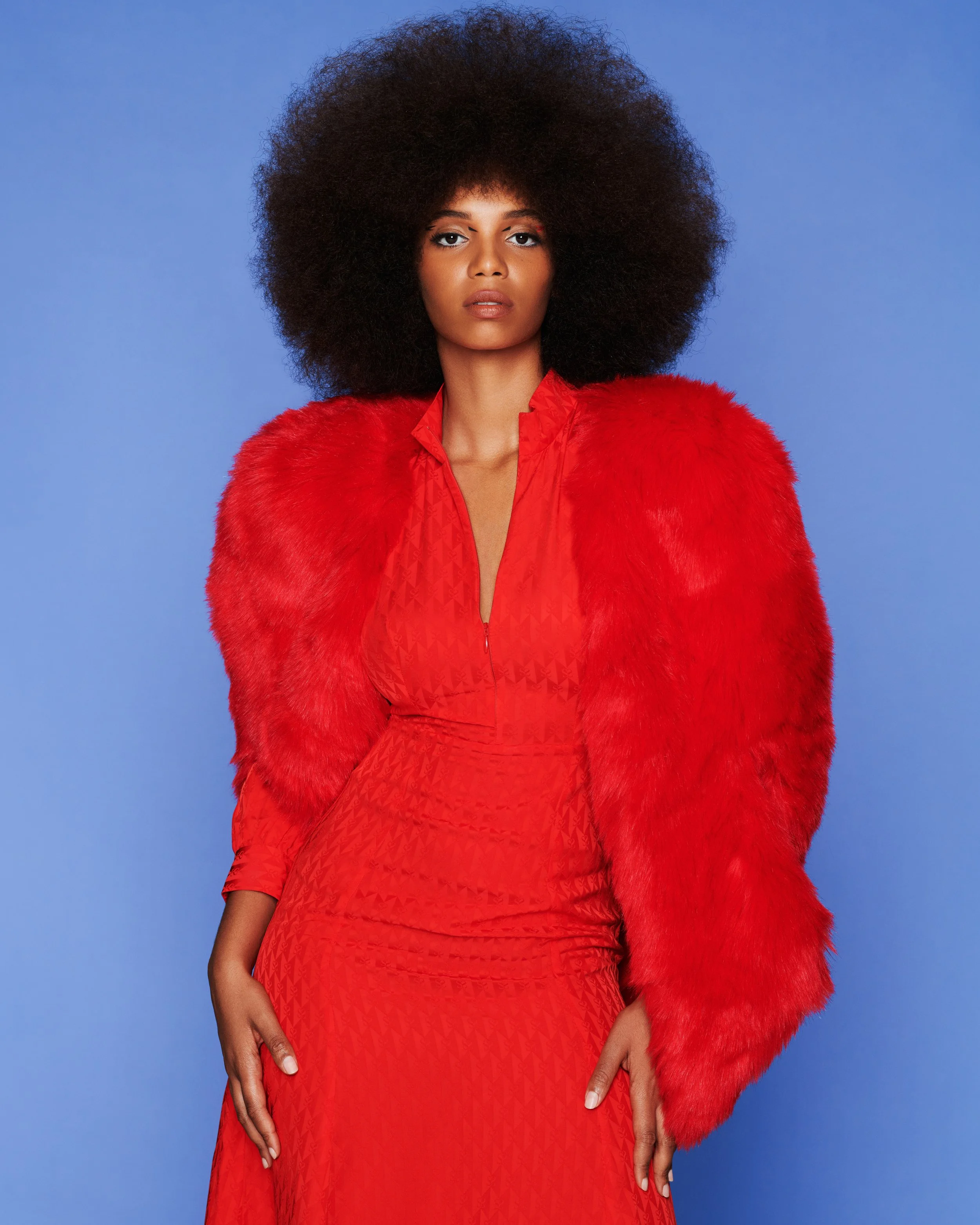 A woman with large, natural curly hair is wearing a red dress and a red faux fur coat, standing against a blue background.
