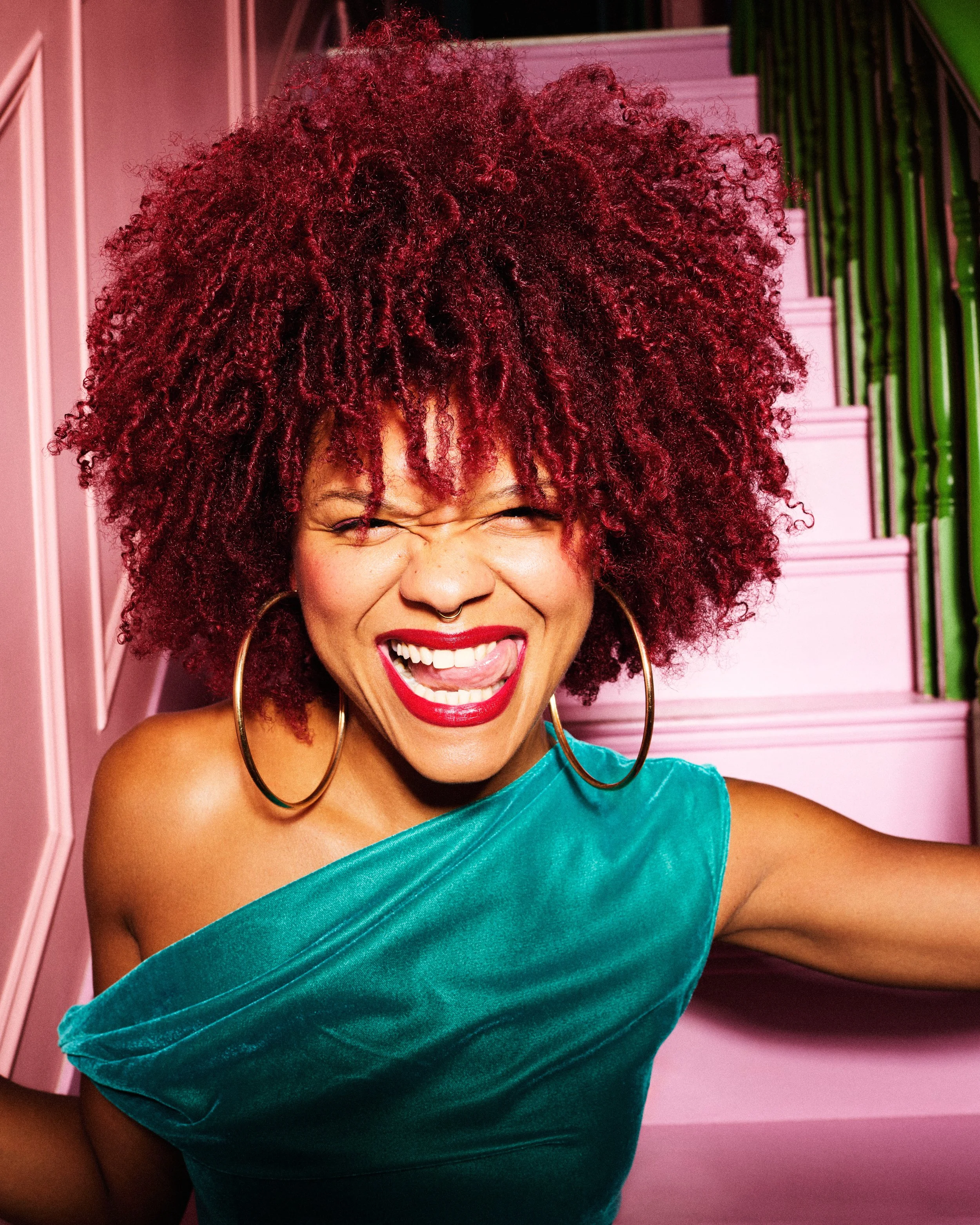 Close-up of a woman with vibrant, curly red hair, wearing a teal velvet dress and large gold hoop earrings, smiling with her eyes closed in front of pink and green background.