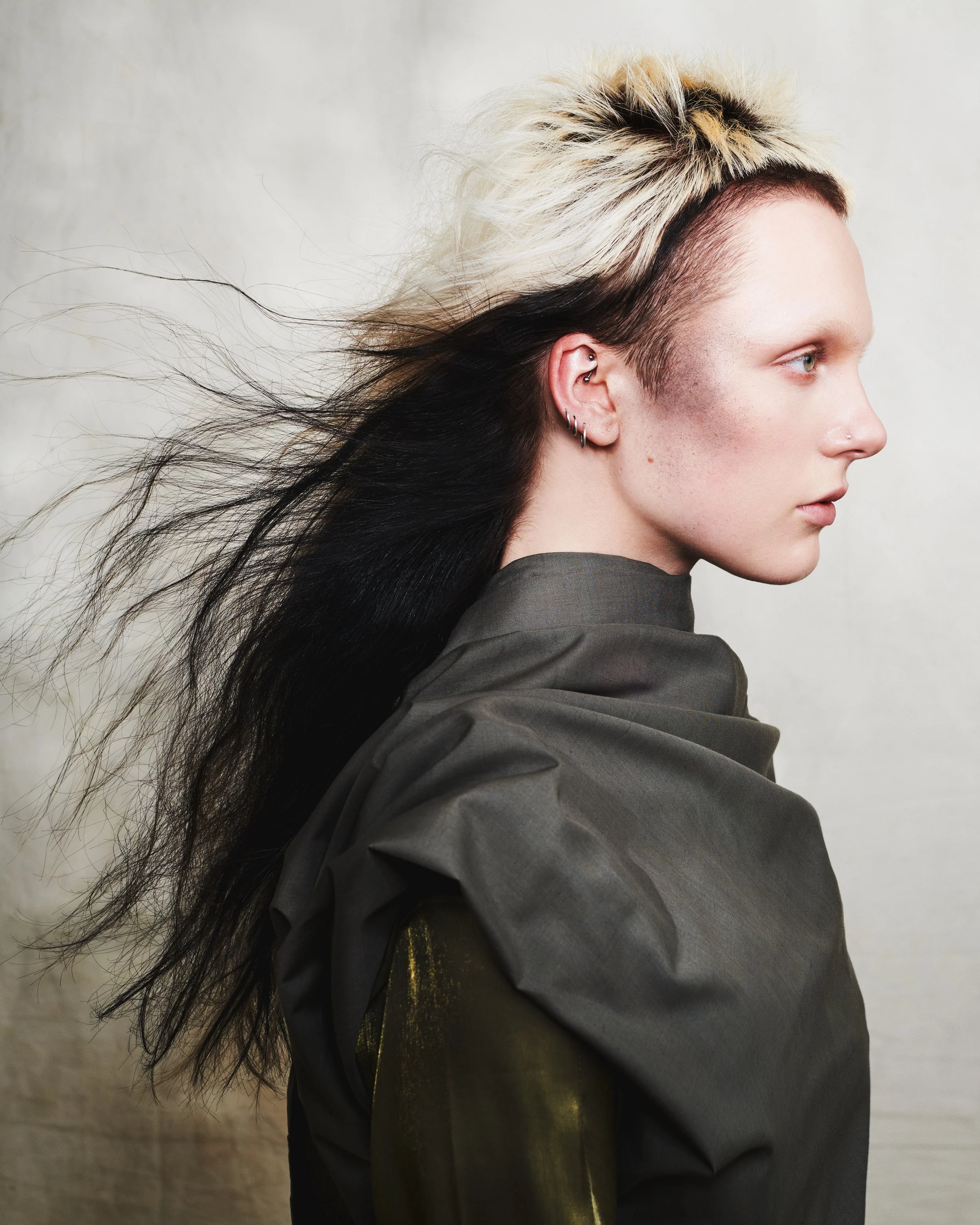 Side profile of a person with light skin, platinum blonde and black hair flowing to the left, wearing multiple piercings in the ear, with a nose piercing, wearing a dark gray or black garment, against a plain white background.