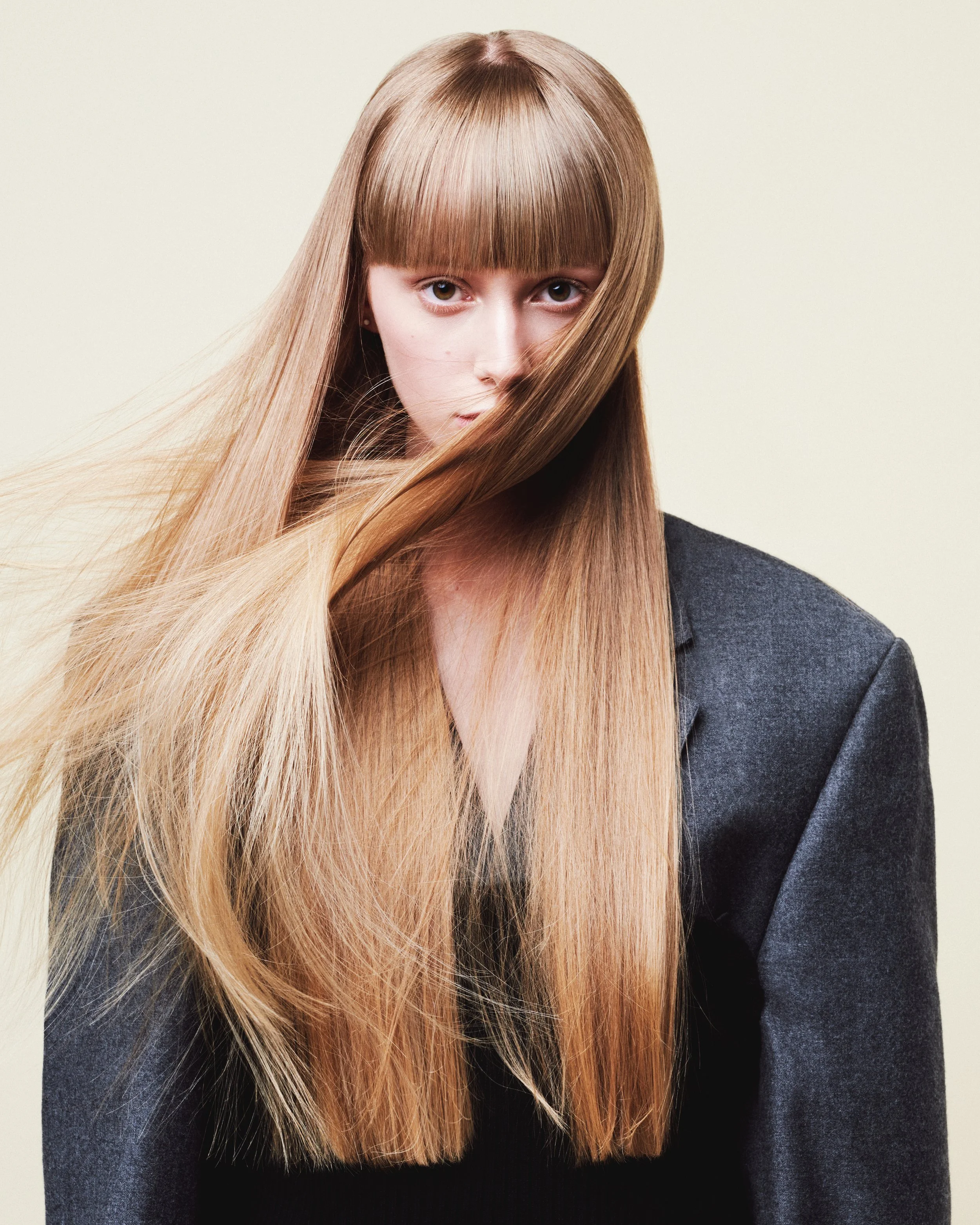 A woman with long, straight, blonde hair with bangs. She is wearing a dark gray blazer against a light background.