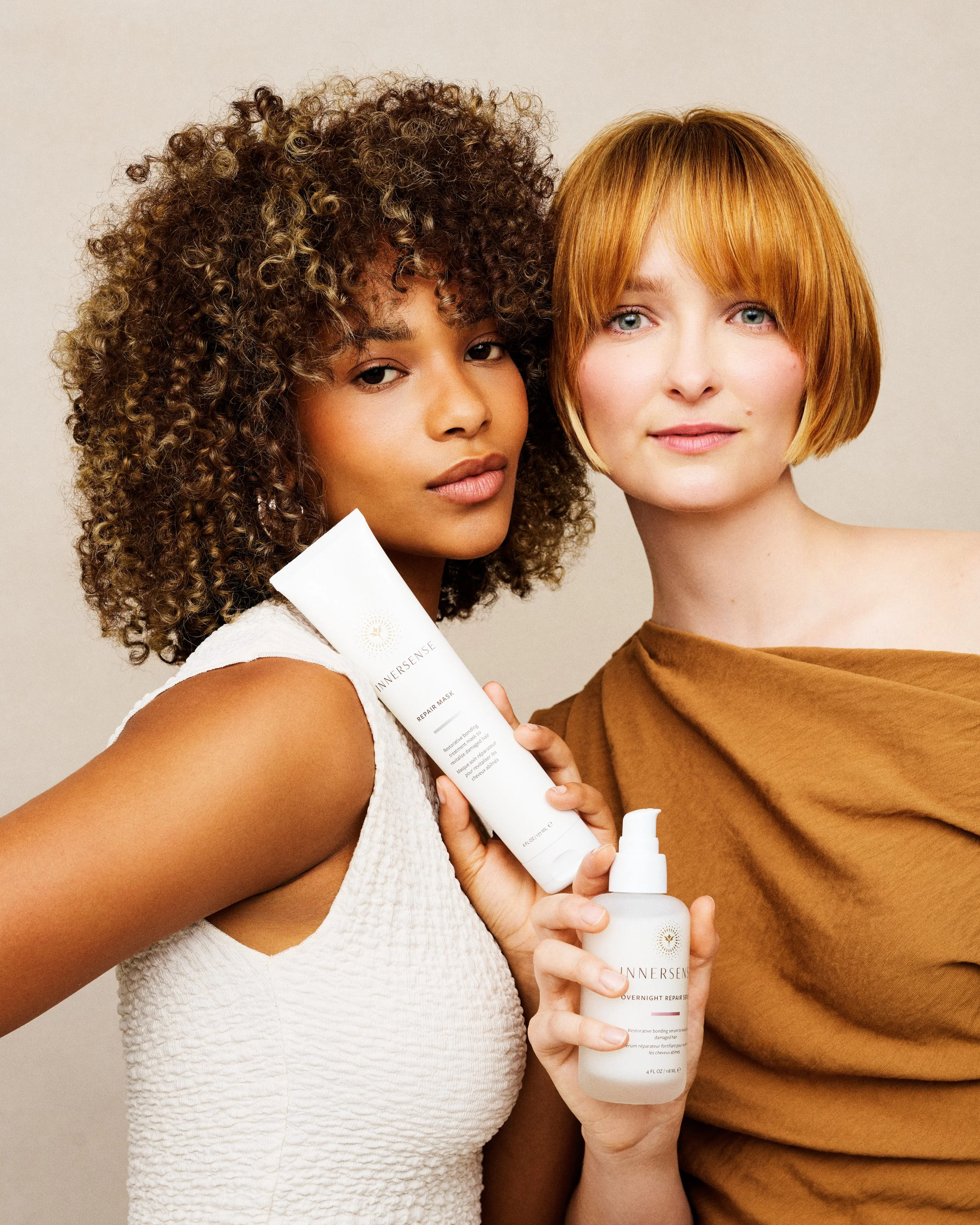 Two women with different skin tones and hair colors holding skincare products, against a plain background, facing the camera.