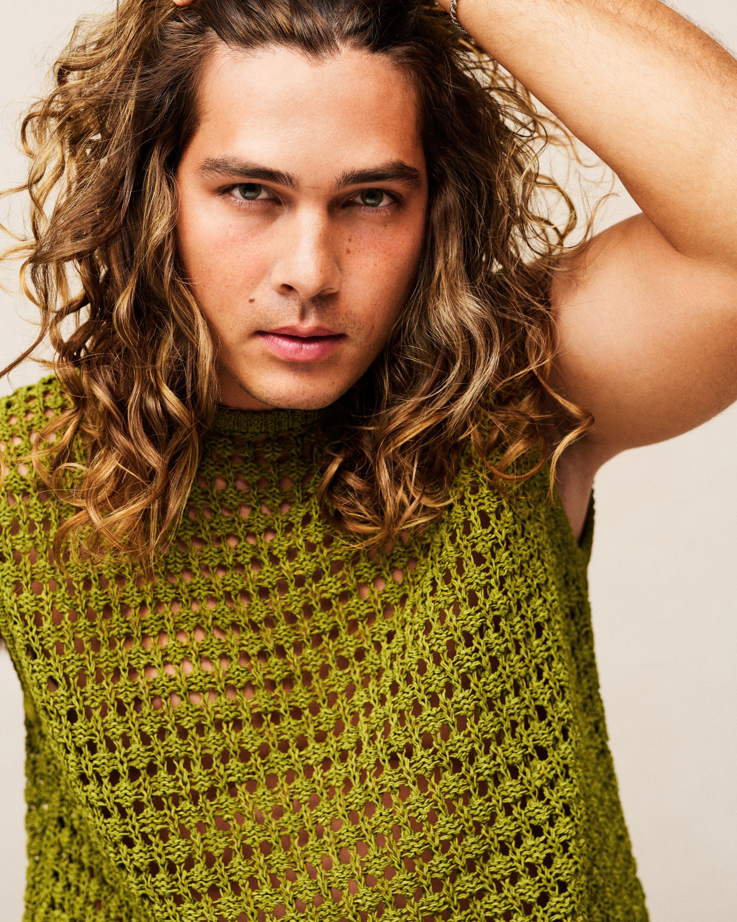 Close-up of a young man with long, curly blonde hair, wearing a green crochet top, posing with his left arm raised and resting on his head.