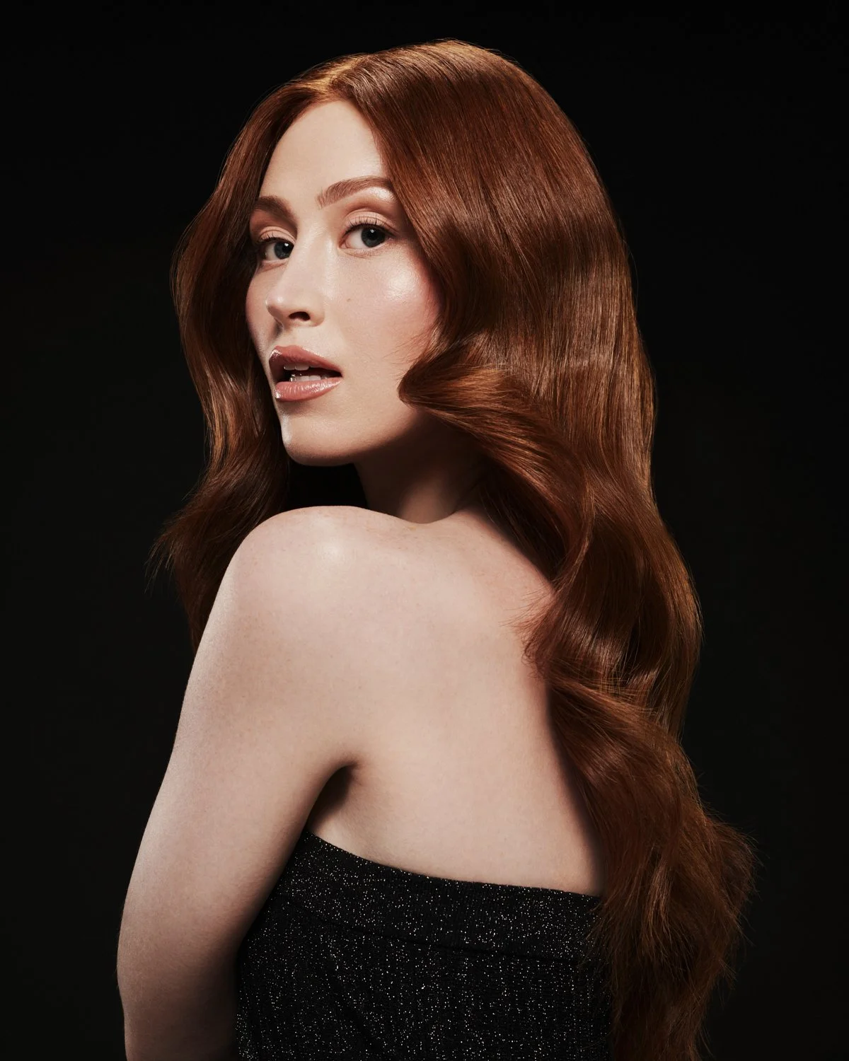 A woman with long, wavy red hair looking over her shoulder against a black background.