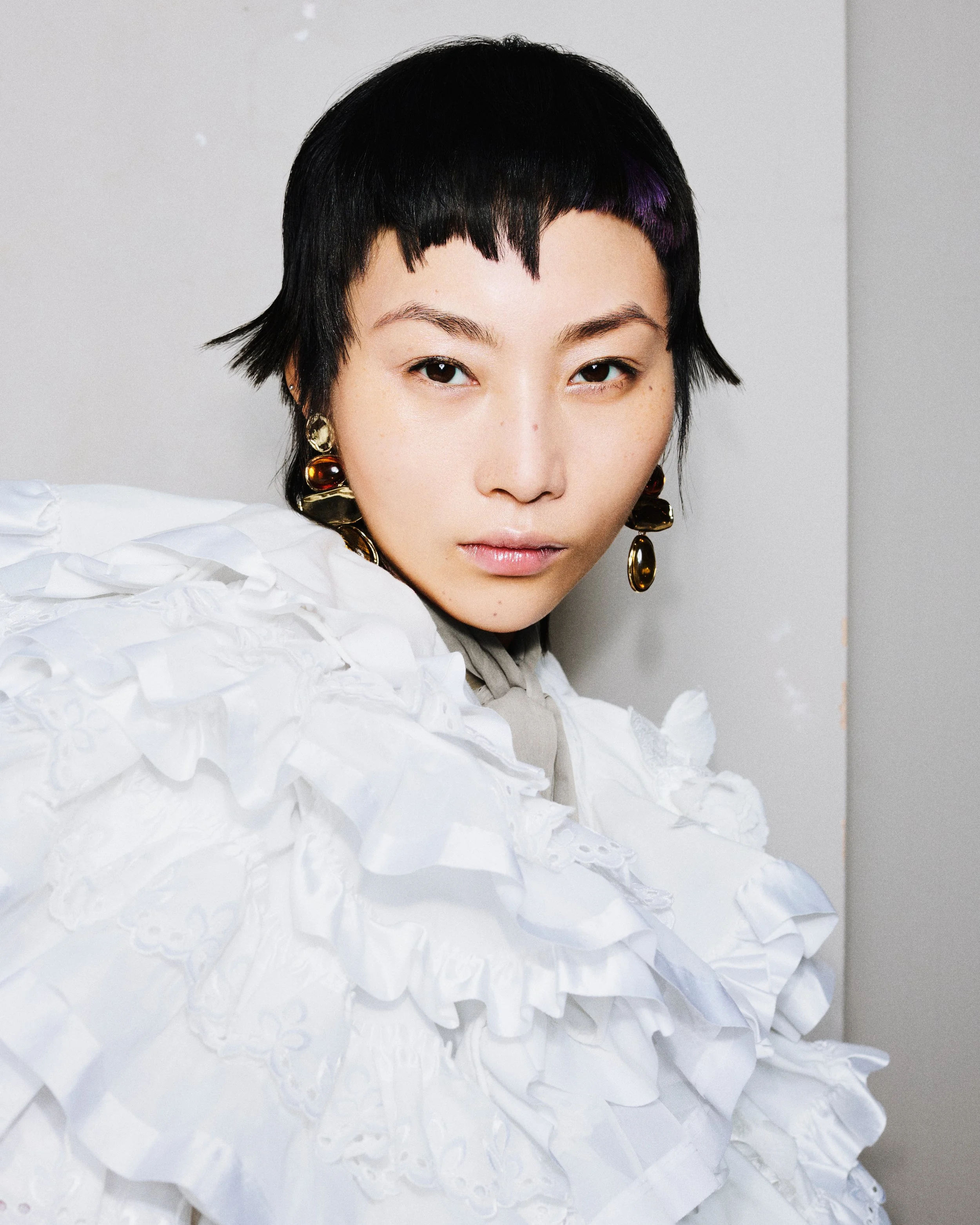 A close-up of a woman with short black hair, wearing large, shiny gold earrings, and a white textured outfit, looking directly at the camera.