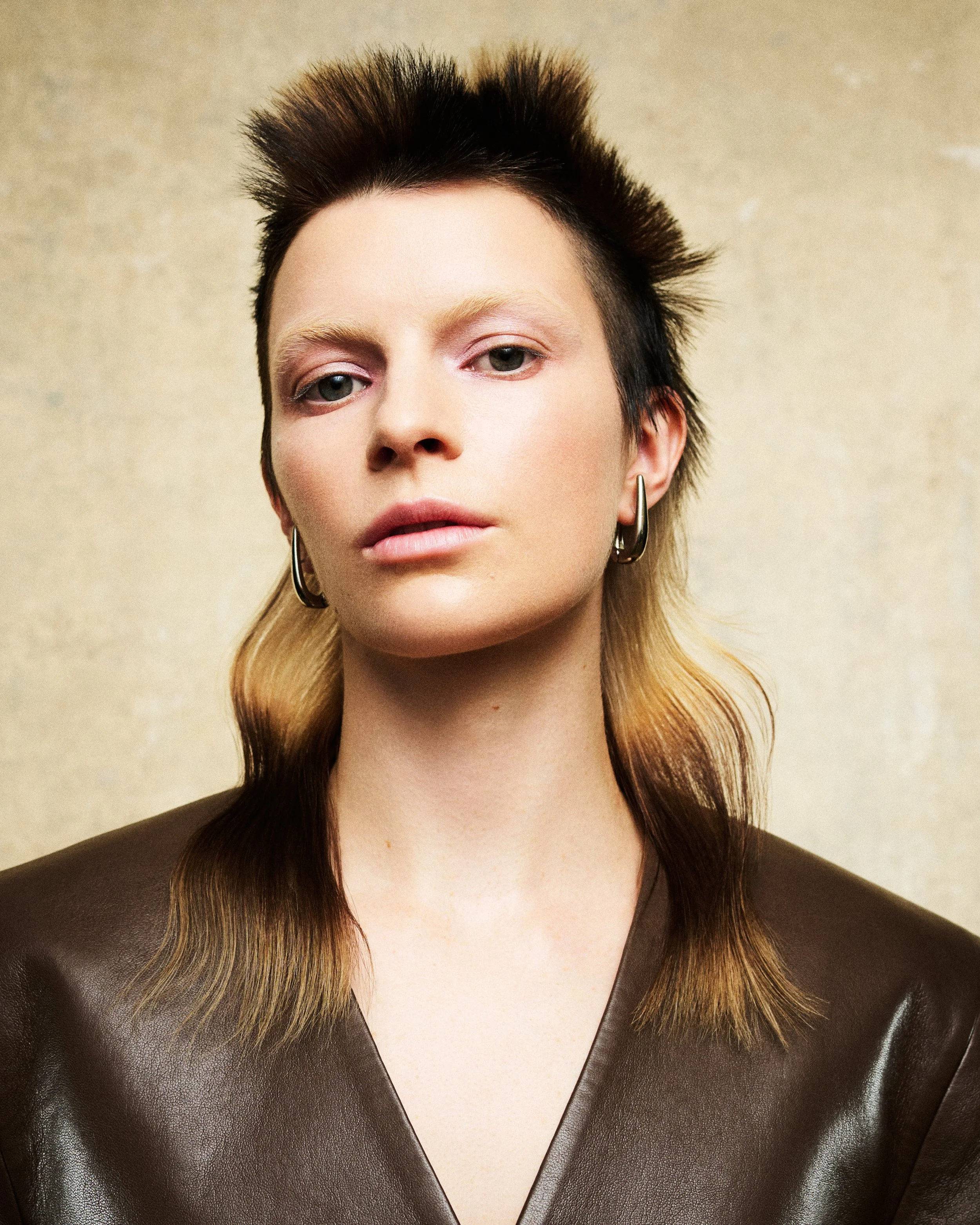 Close-up of a woman's face with short, spiked hair, wearing hoop earrings, and a brown leather top, against a neutral background.