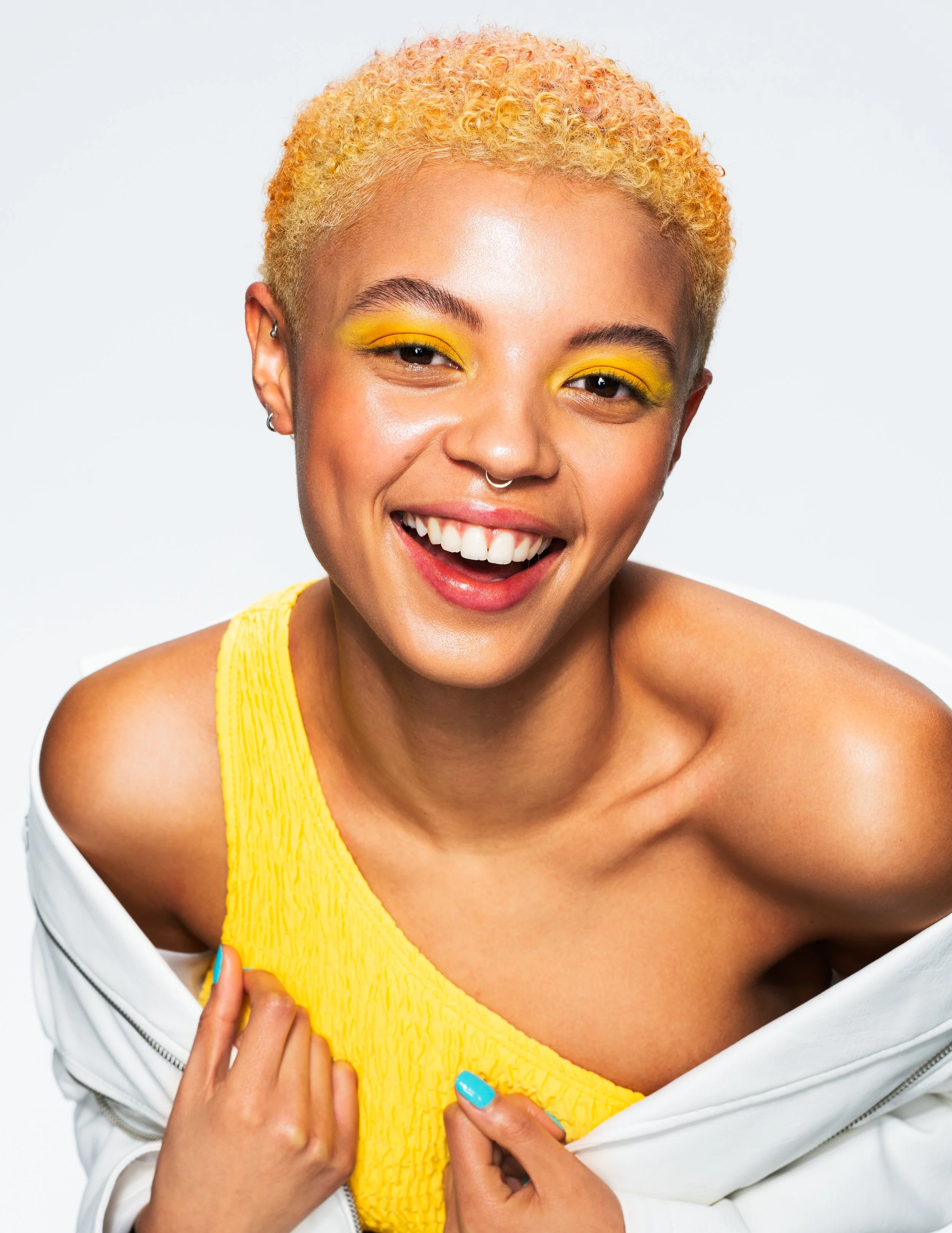 Close-up of a smiling woman with yellow curly hair, wearing bright yellow eyeshadow, a yellow top, and a white jacket, against a neutral background.