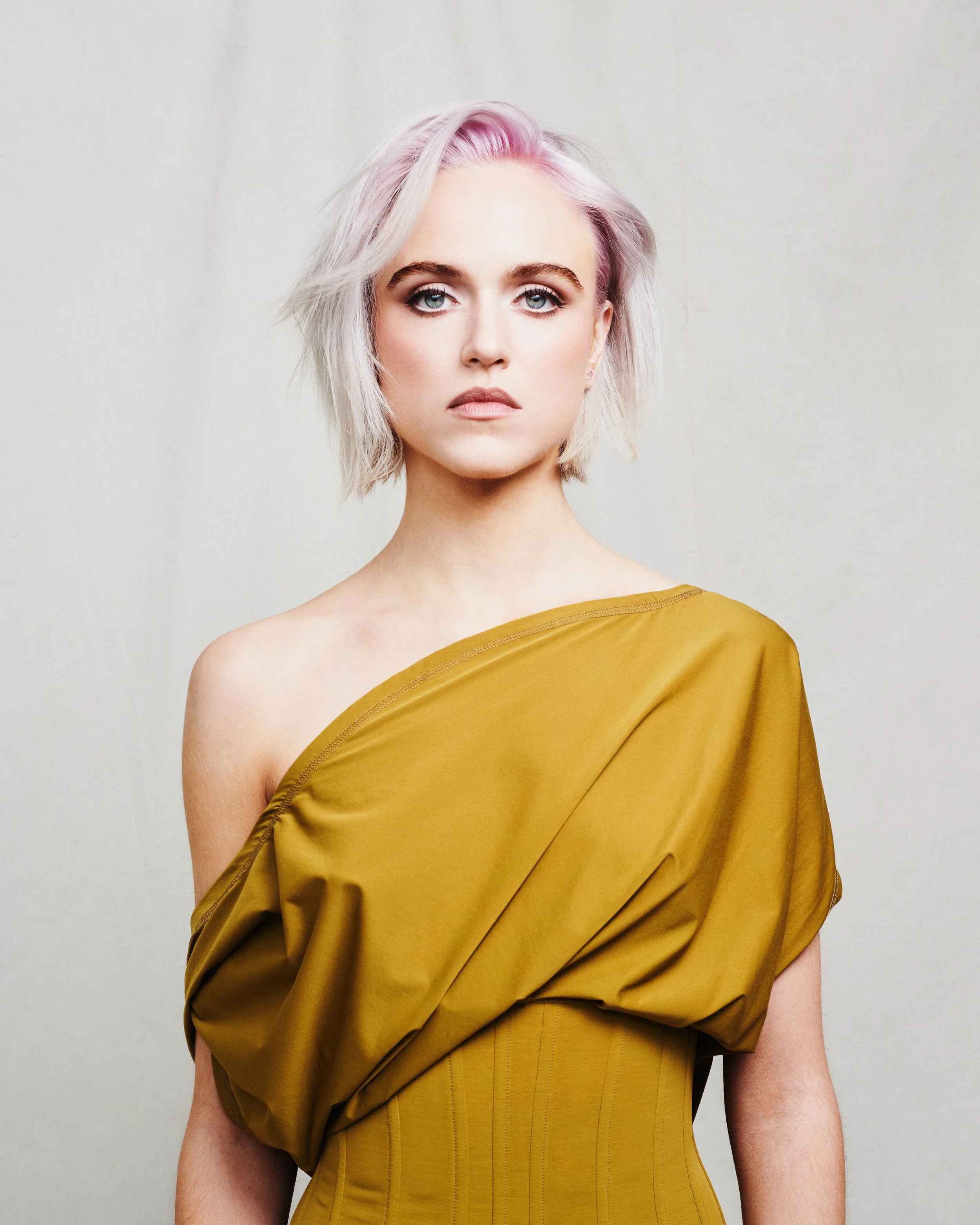 Close-up of a woman with platinum blonde hair with light pink highlights, wearing a yellow off-shoulder dress, standing against a plain light grey background.