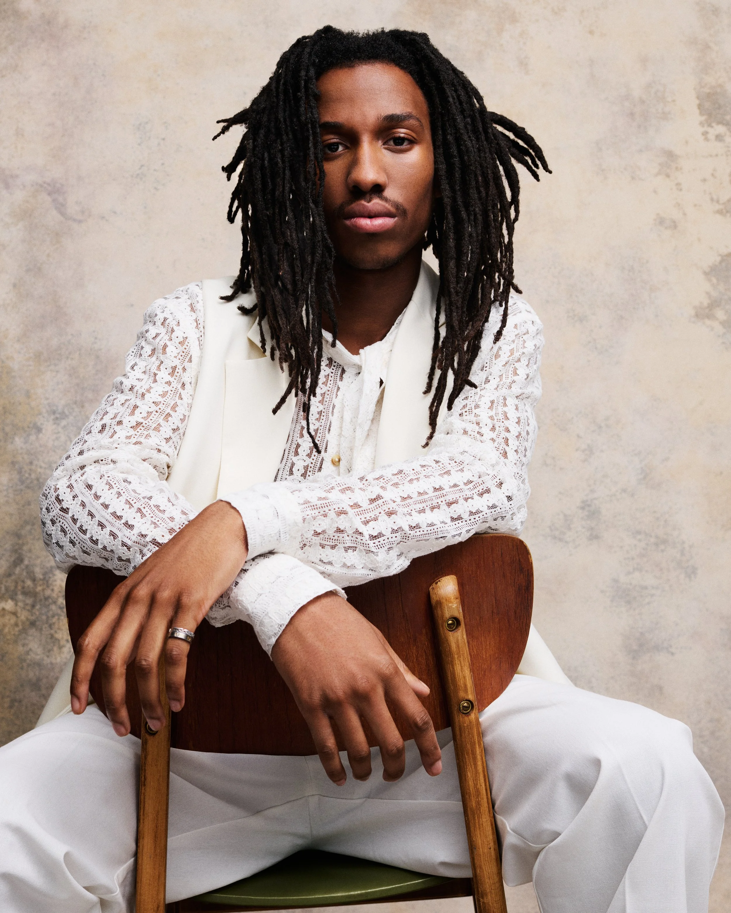 A person with black dreadlocks wearing a white lace shirt and white pants, sitting on a wooden chair with their arms crossed over the backrest, against a neutral beige wall.