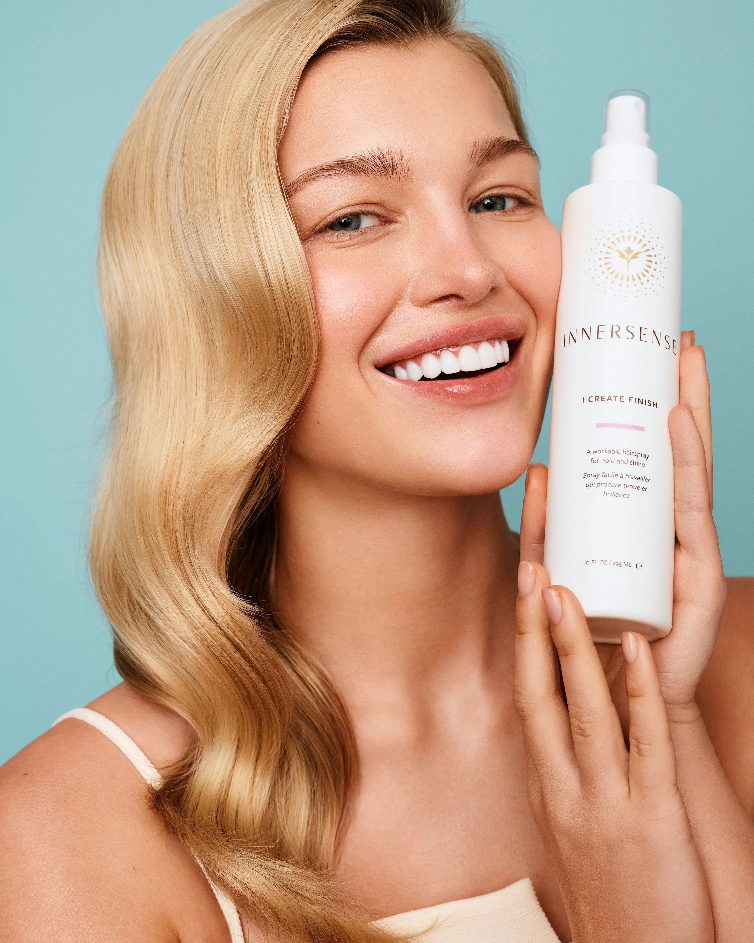 A woman with long blonde hair smiling and holding a white bottle of InnerSense hair spray with a pastel blue background.