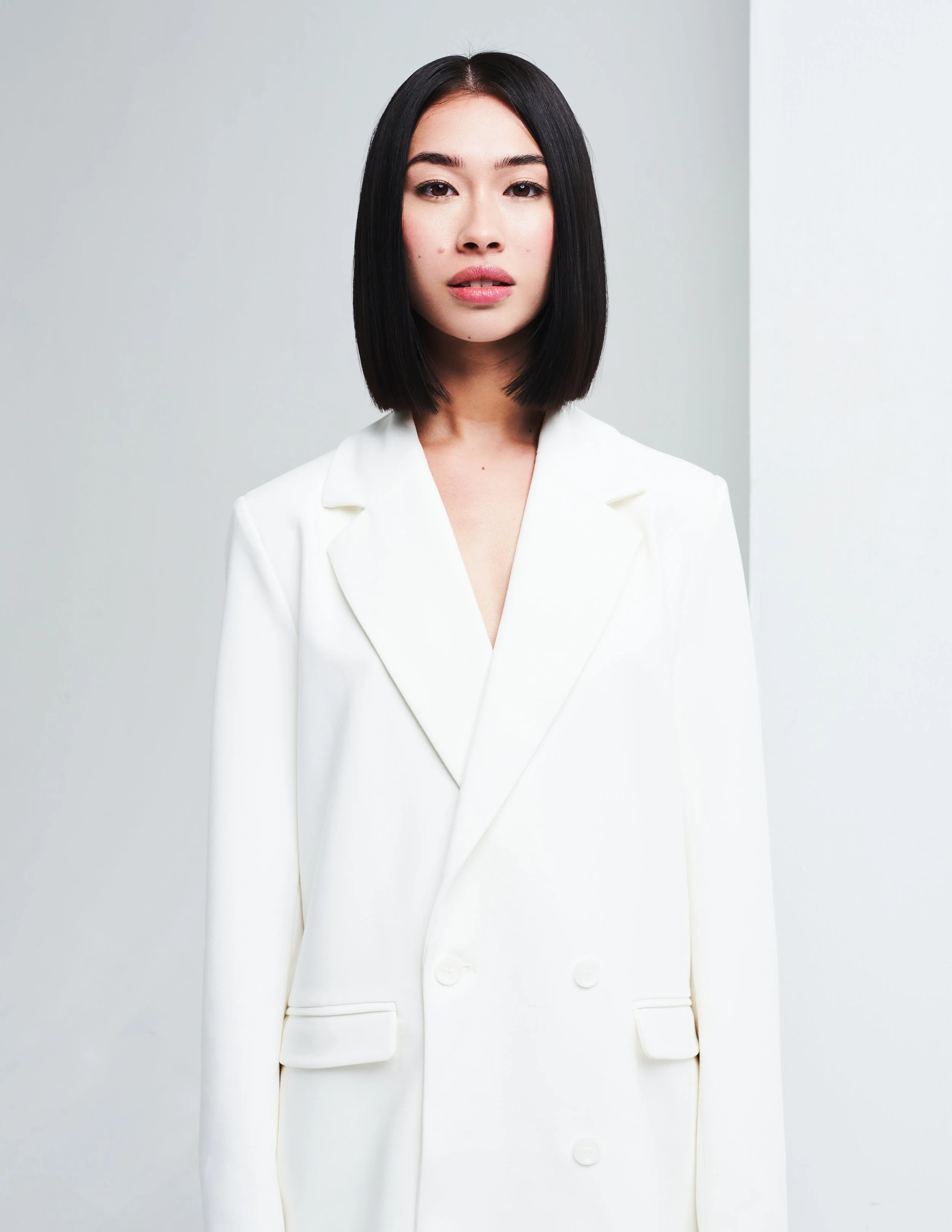 A woman with straight, black shoulder-length hair and light makeup, wearing a white blazer, standing against a light gray background.