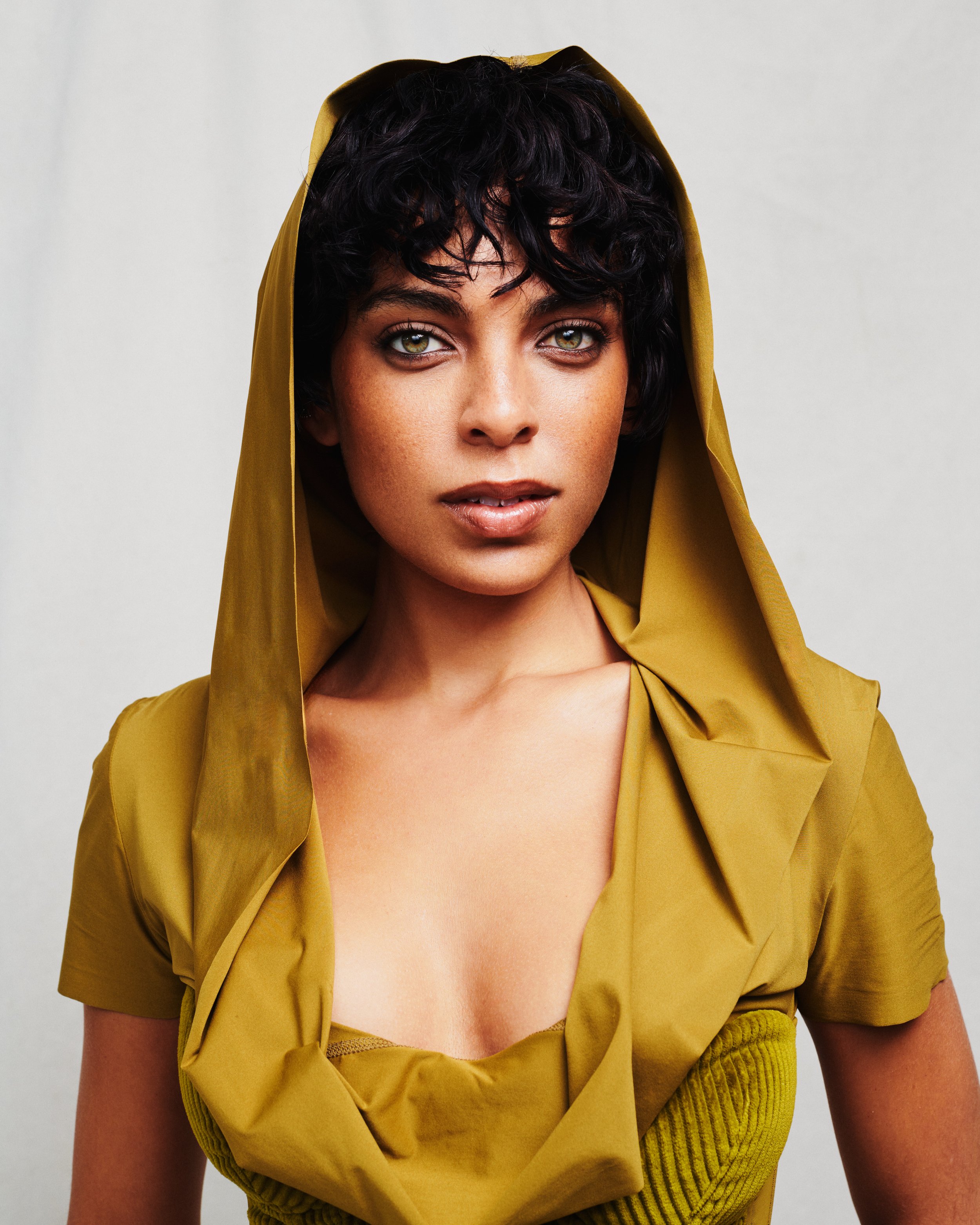 Close-up of a woman with curly dark hair and piercing green eyes, wearing a yellow garment with a hood, against a plain light background.