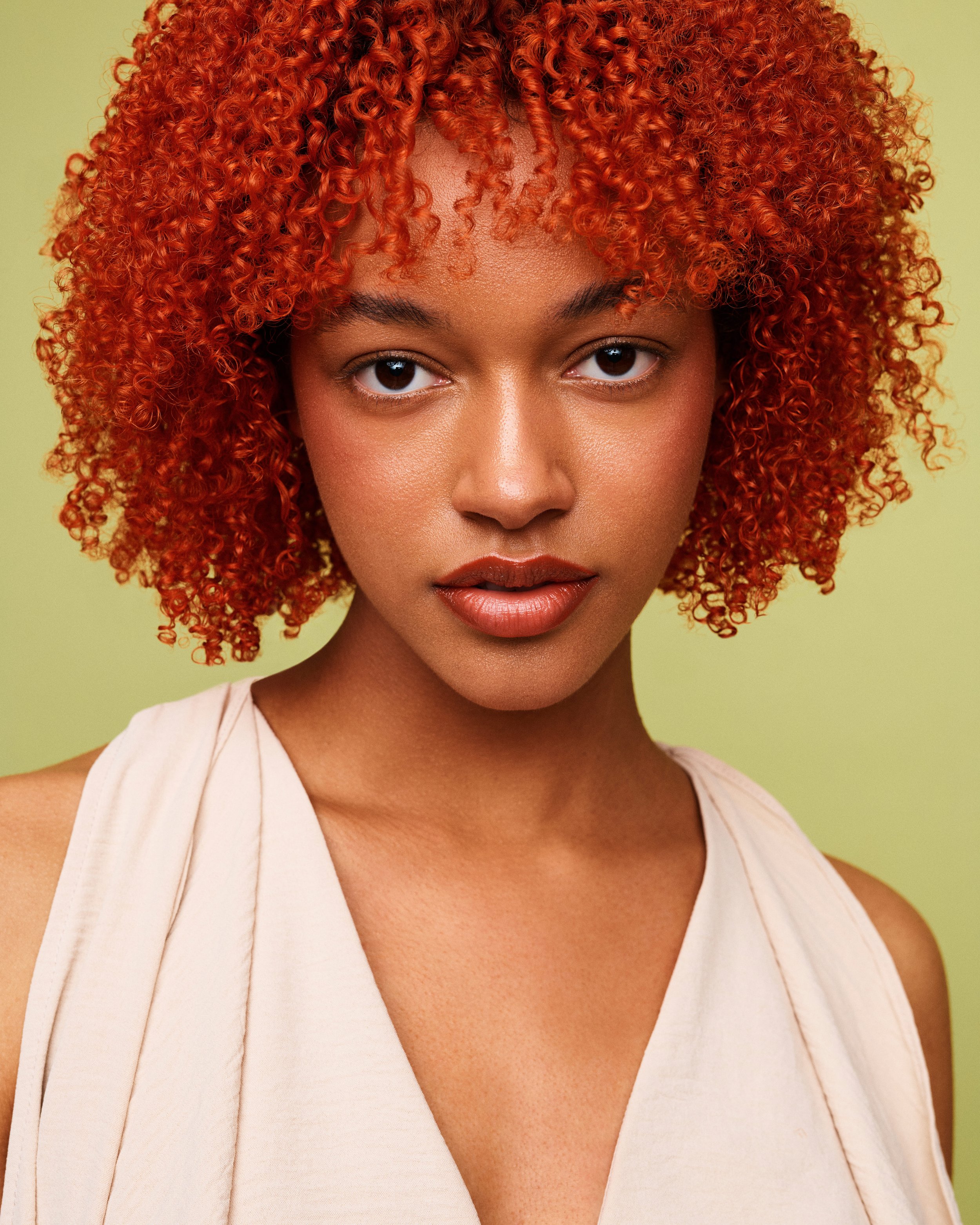 Close-up of a woman with red, curly hair, wearing a light-colored top, against a light green background.