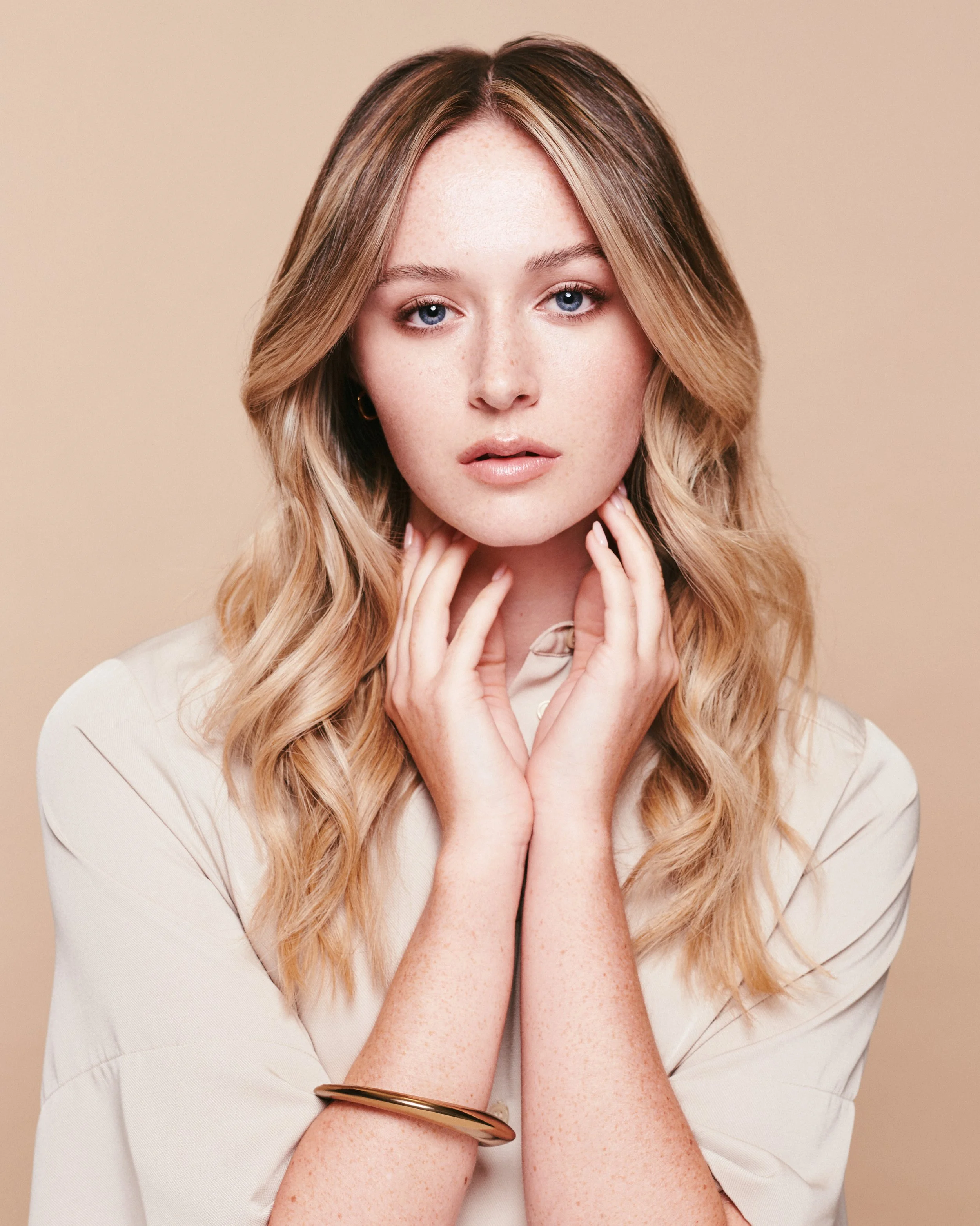 A woman with long, wavy blonde hair and blue eyes, wearing a beige blouse and gold jewelry, looking directly at the camera with her hands gently touching her neck and face.