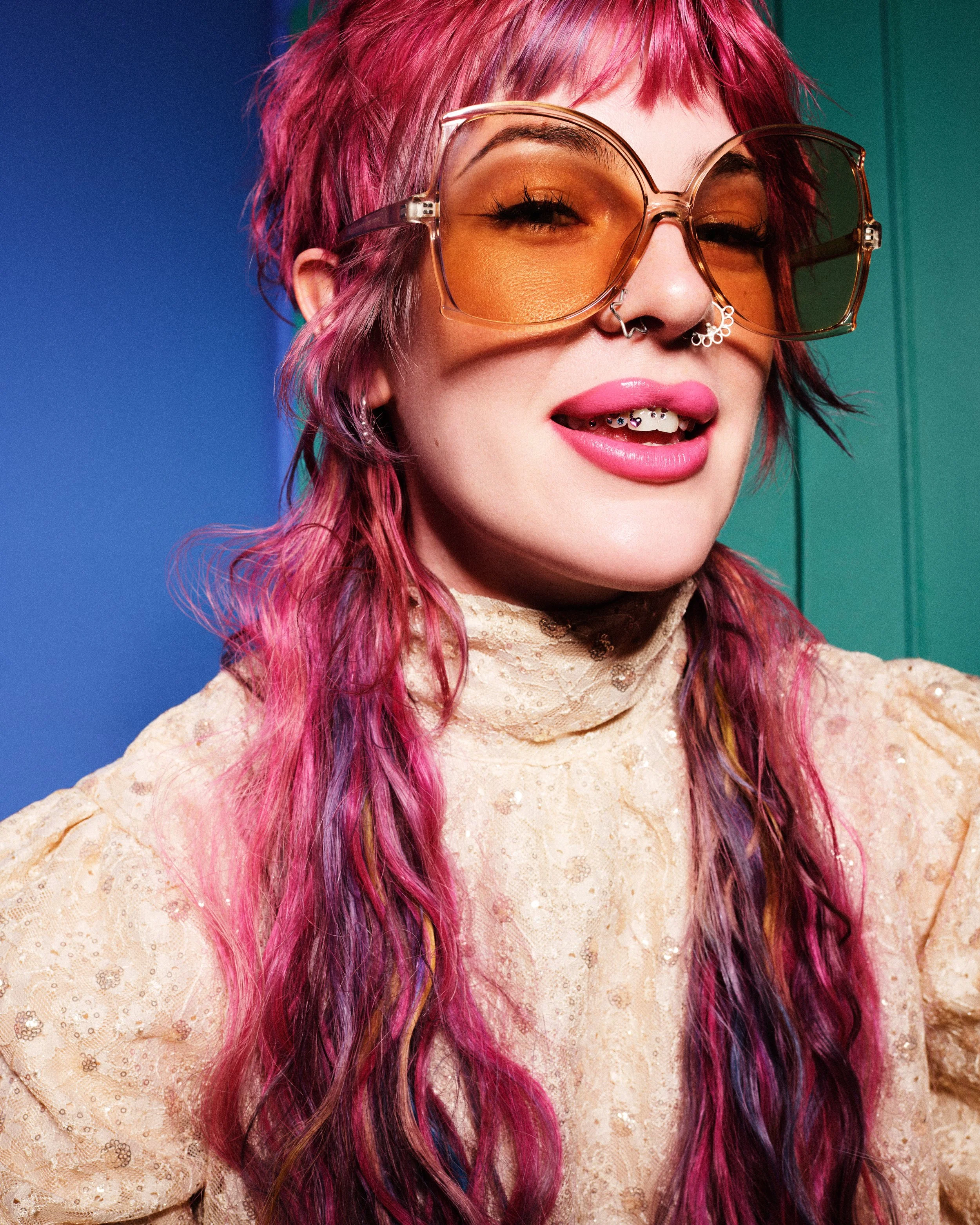 A person with vibrant pink hair, wearing oversized brown sunglasses and pink lipstick, smiling with visible braces. The person is dressed in a beige lace top, standing against a background divided into blue and green sections.