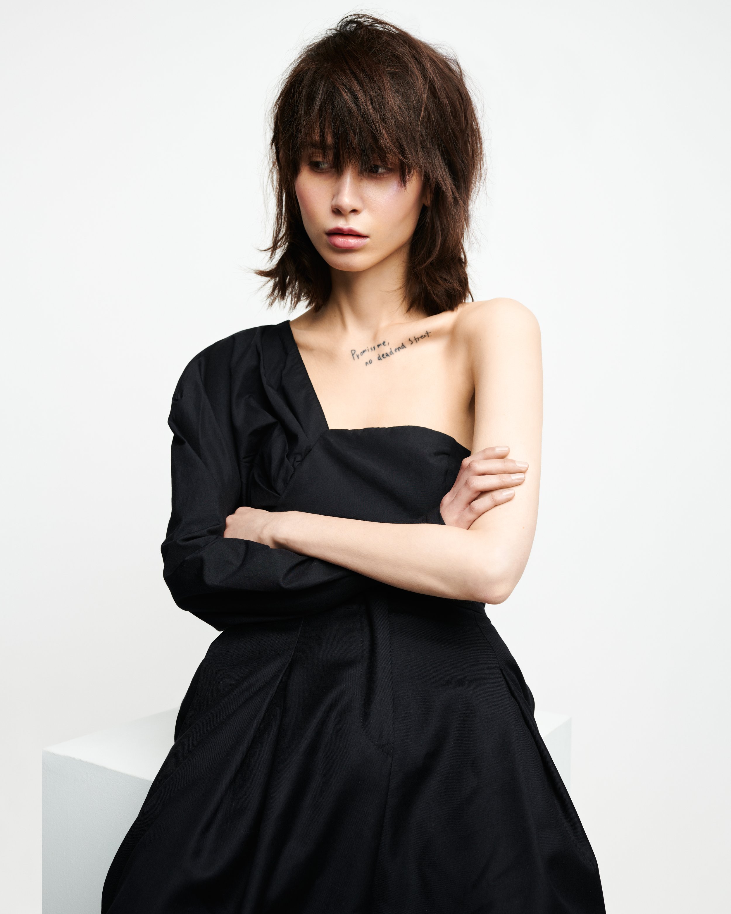 A woman with short, shaggy brown hair, wearing a black off-shoulder top, standing against a plain white background, with a tattoo on her chest that says 'Promise me no dead end street.'