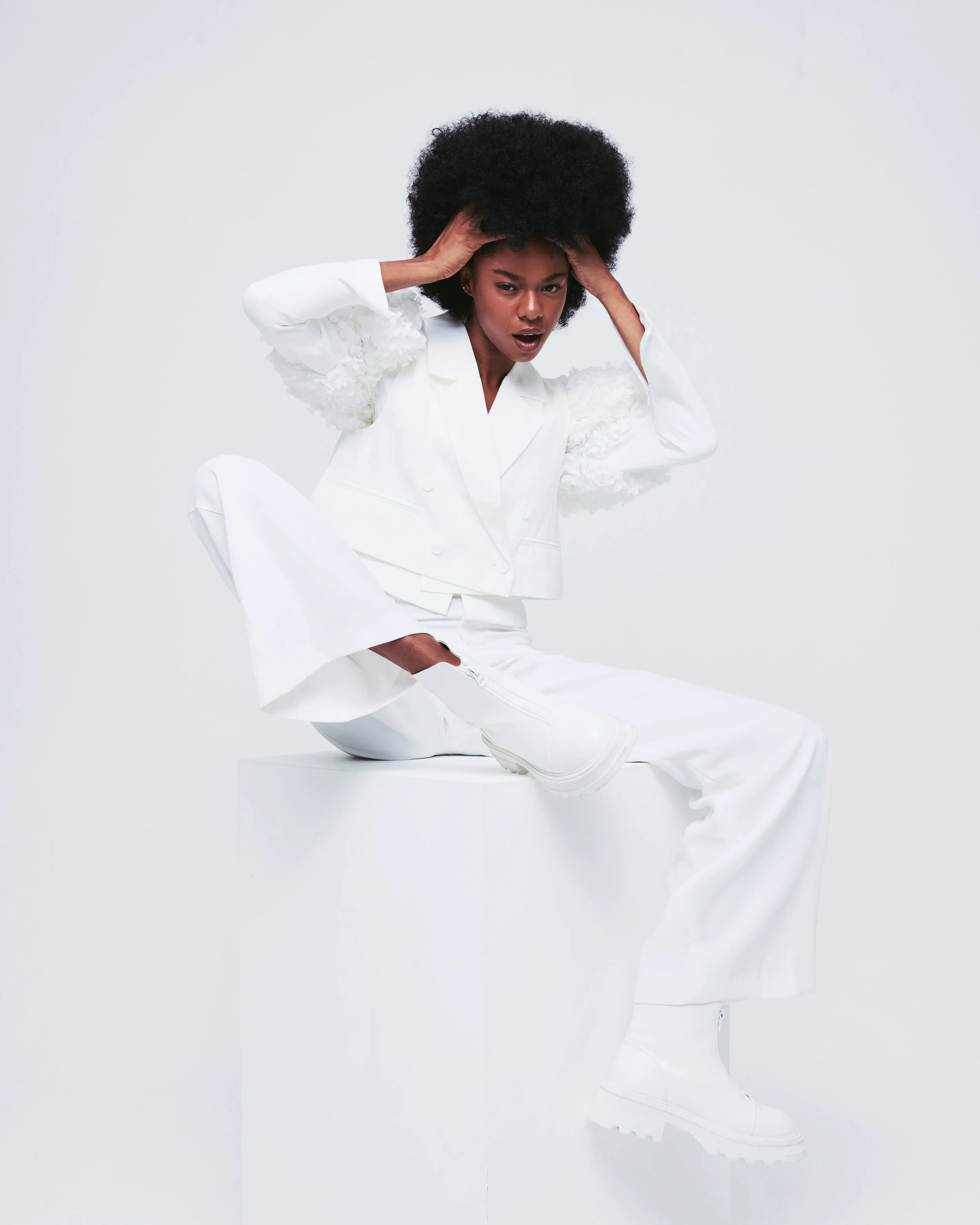 A woman dressed in all white, posing against a white background. She is crouching on a white pedestal, with one leg bent and the other extended, wearing white clothing and chunky white boots. She has natural curly hair and is holding her head with bo