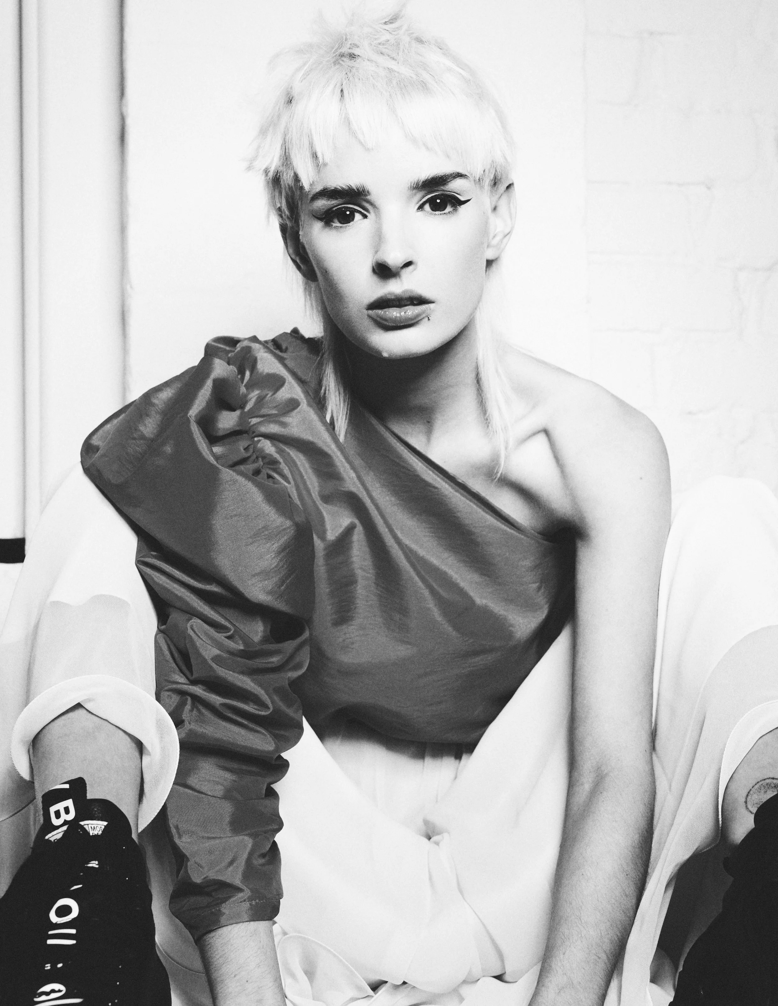 Black and white photo of a woman with short, platinum blonde hair and makeup, sitting on a white surface, wearing a dark, textured top and led Nike sneakers.