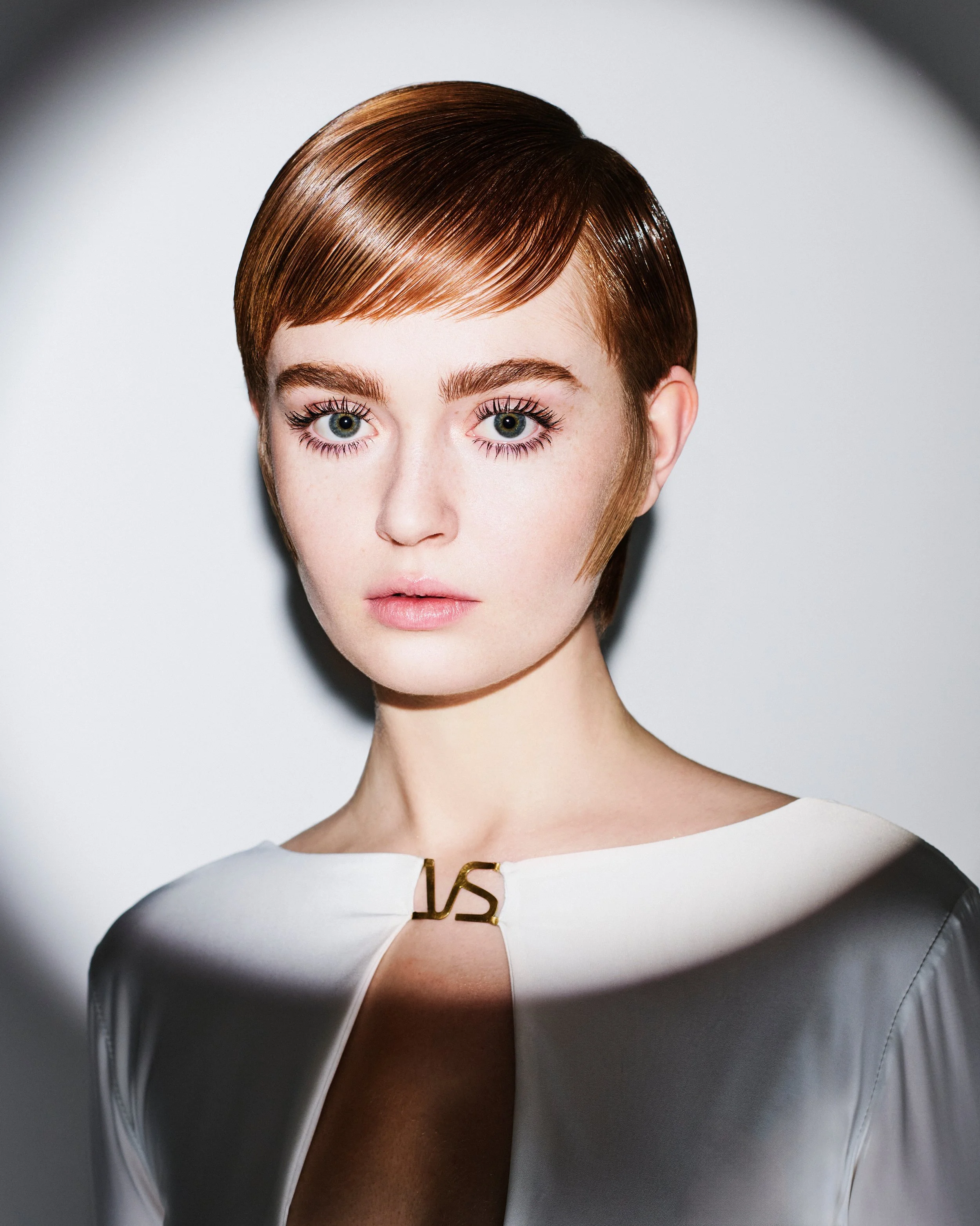 Close-up of a woman with short, sleek, reddish-brown hair, wearing a white dress with a gold zipper detail at the neckline, standing against a plain white background.