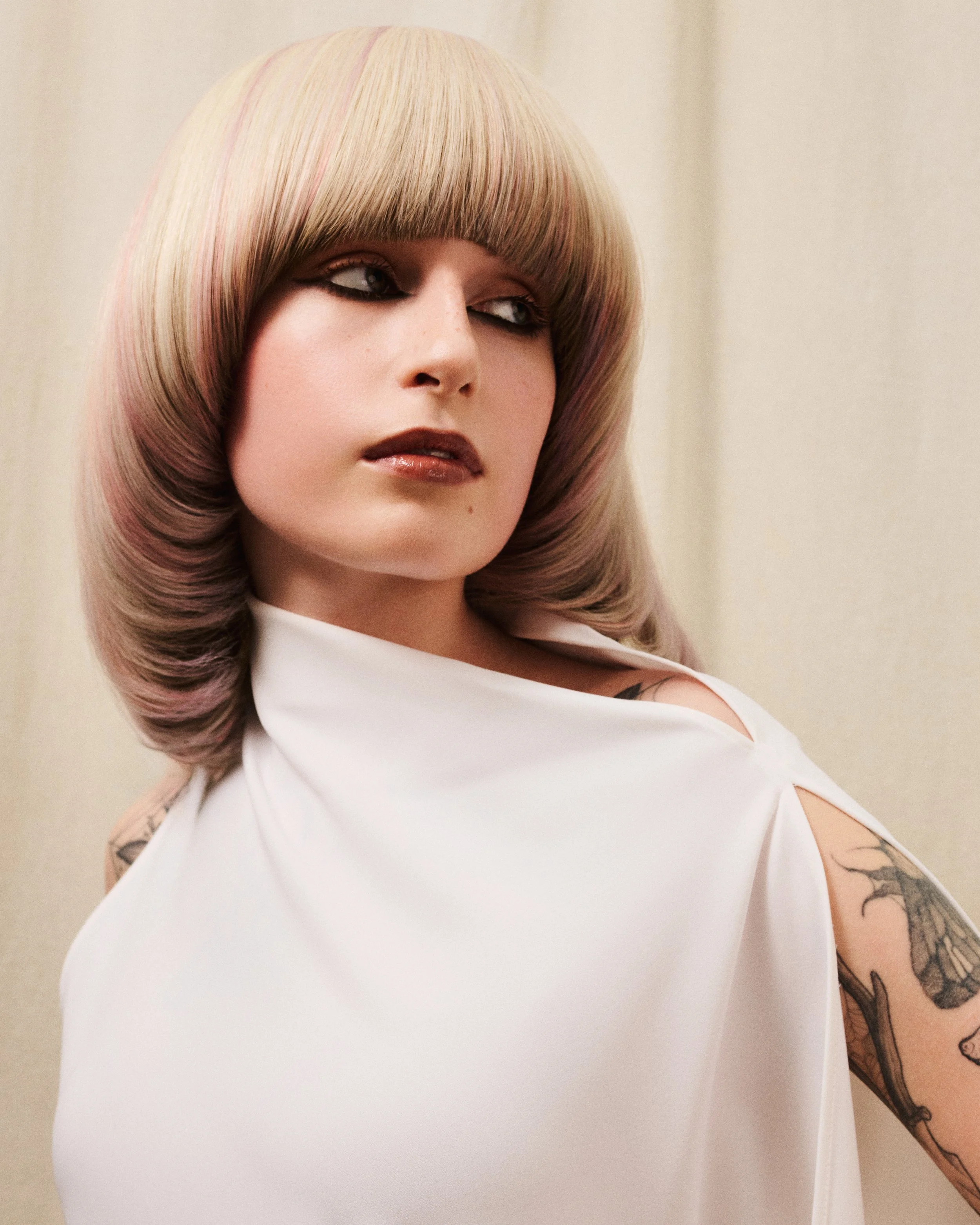 A woman with blonde hair styled in a voluminous, rounded haircut with bangs, wearing a white top with a cut-out shoulder design, and visible tattoos on her arm.