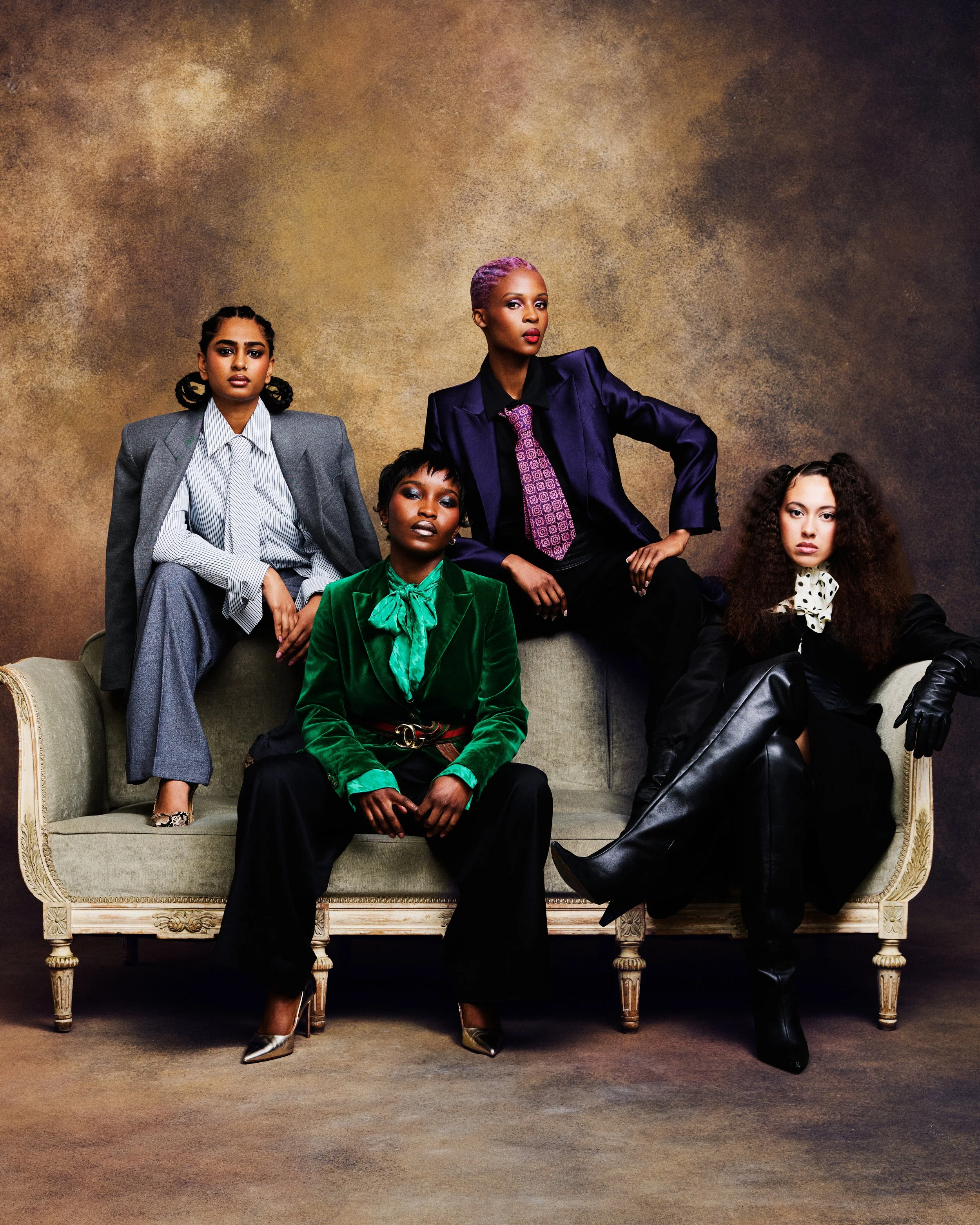 Group of five women dressed in fashionable clothing, posing on and around an ornate vintage sofa against a textured brown background. They have diverse hairstyles and expressions, exuding confidence and style.