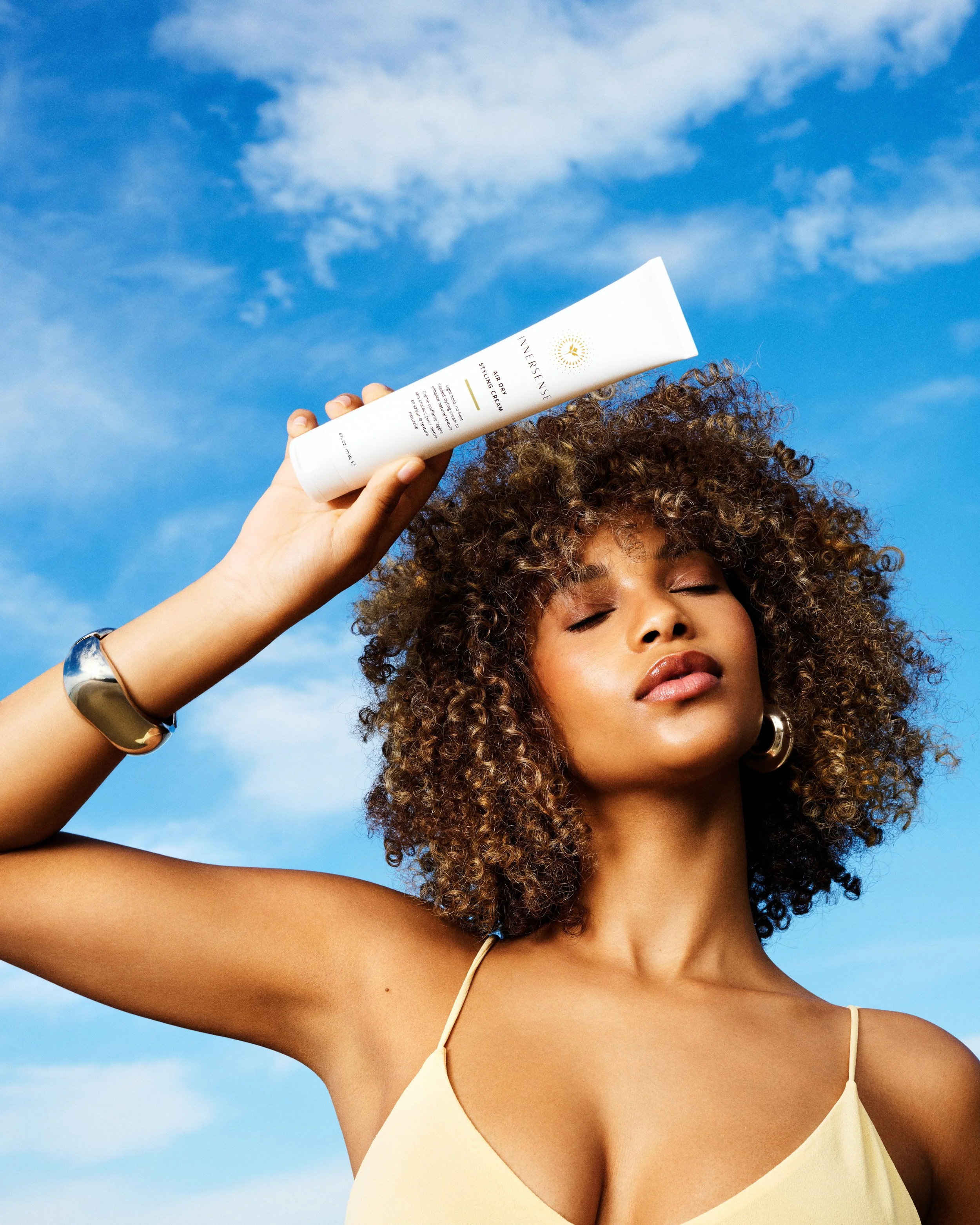A woman with curly hair is holding a tube of Innersense AirRecall Styling Cream up to her forehead against a blue sky with clouds.