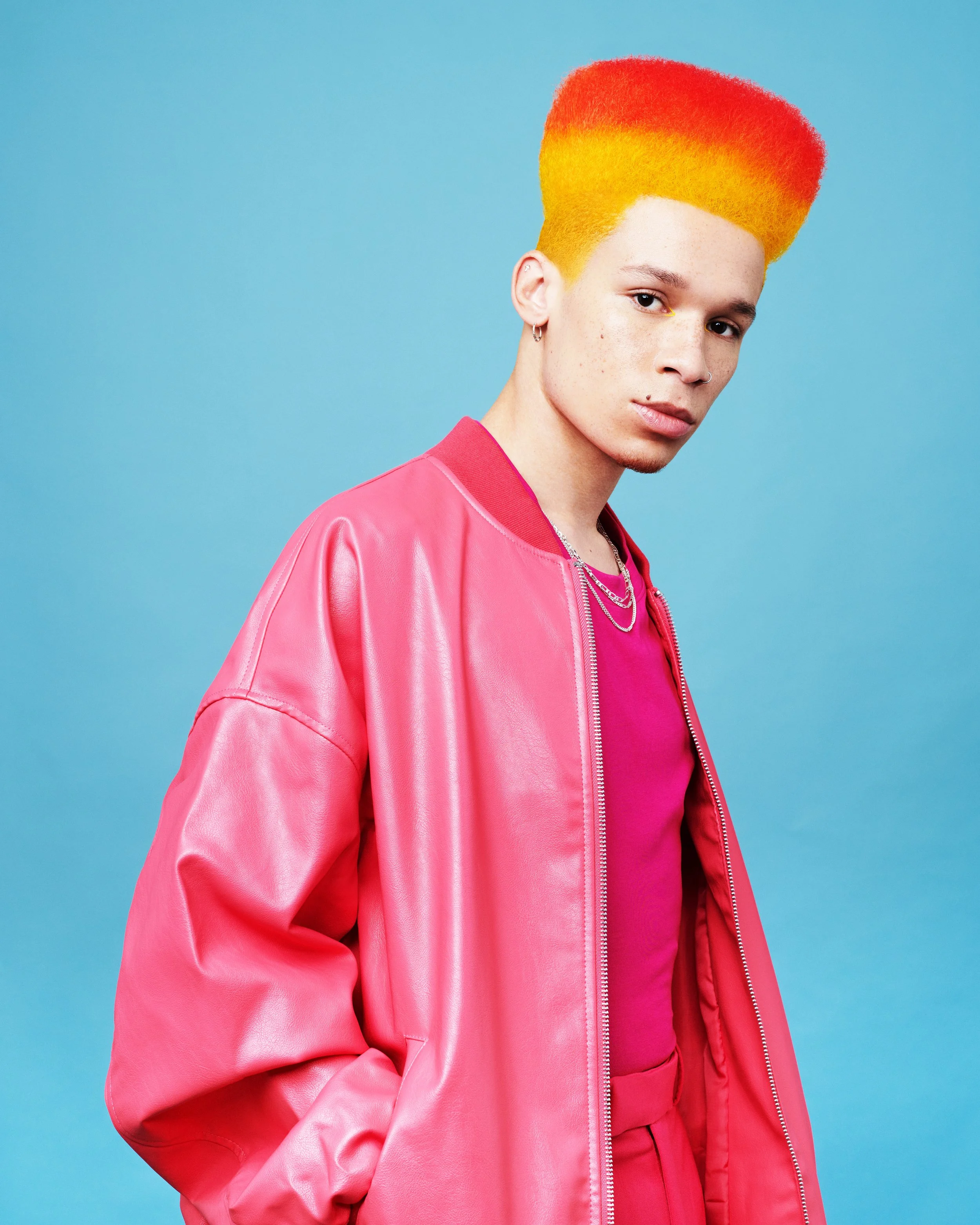 A young person with rainbow-colored hair, fading from red to yellow, wearing earrings and a chain necklace, dressed in a bright pink jacket and matching shirt, standing against a light blue background.