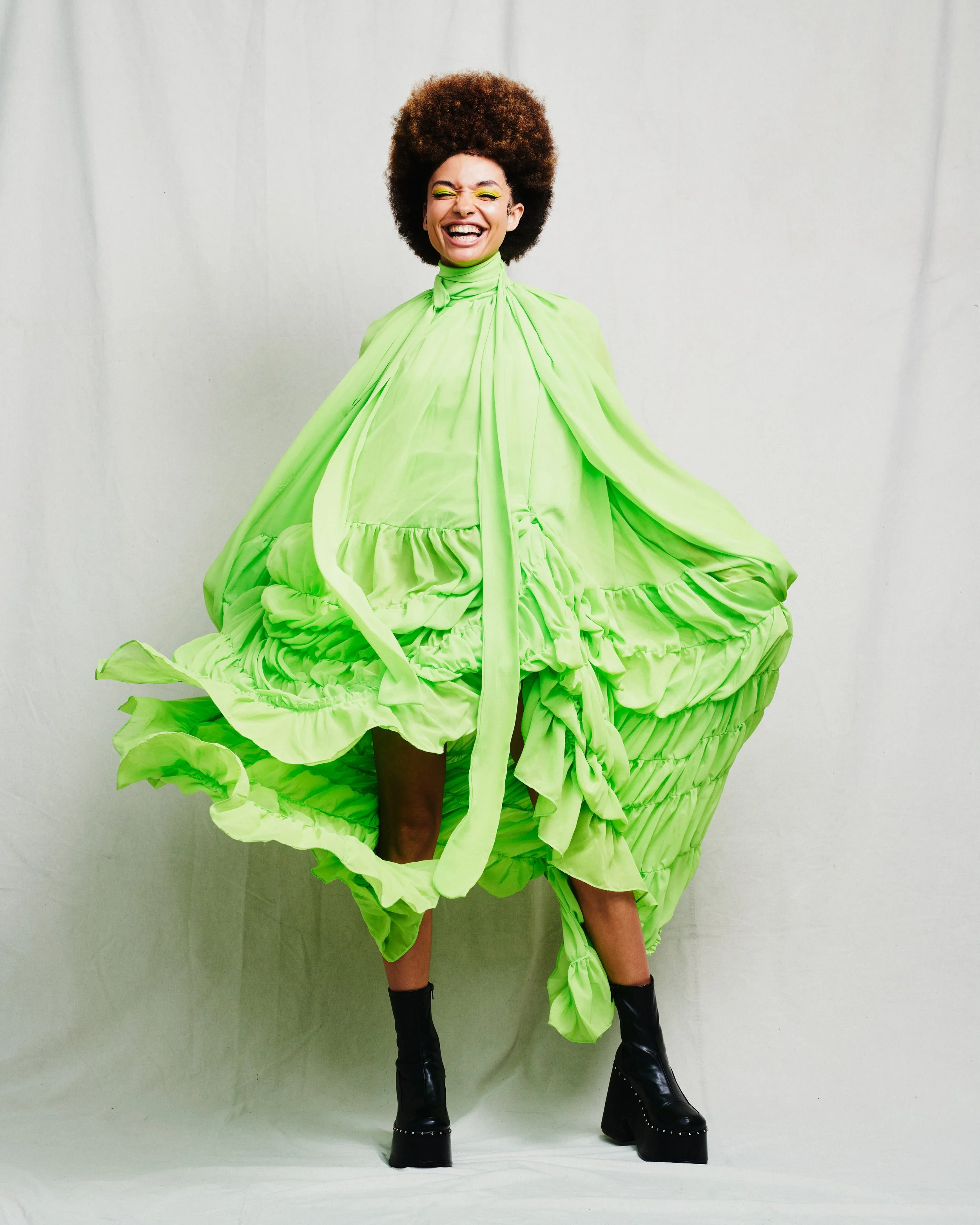 woman wearing a voluminous bright green dress, black platform boots, standing against a plain background, smiling with eyes closed.